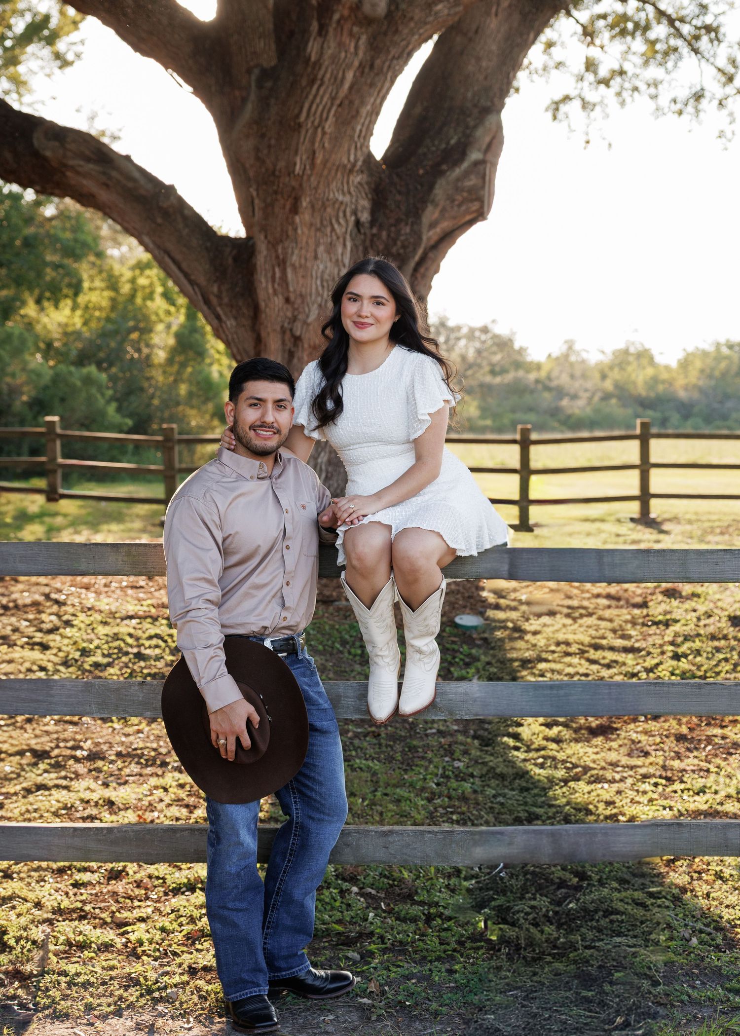 A couple sits together on a rustic wooden fence in a pastoral setting during golden hour.