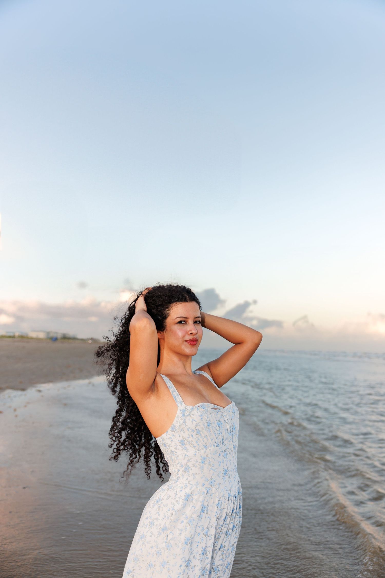 Woman in flowy white dress poses on empty beach at sunset with ocean waves in background and soft lighting.