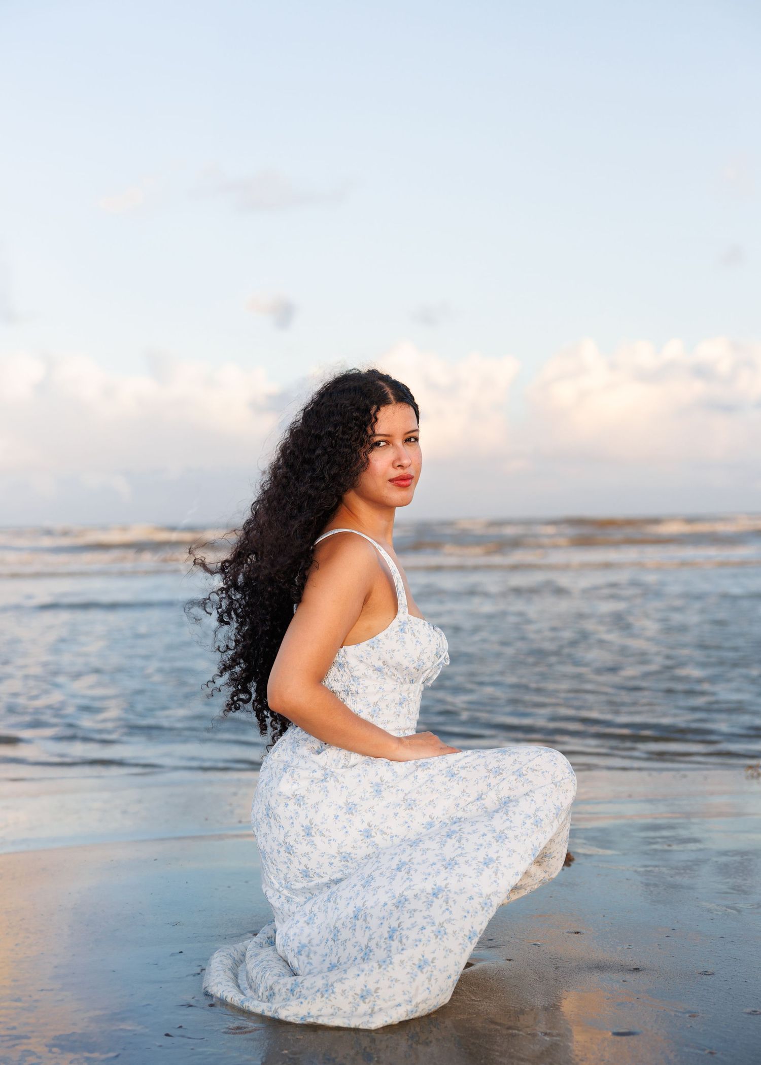 A person in a flowing white dress poses gracefully on a beach at sunset with waves and clouds in the background.