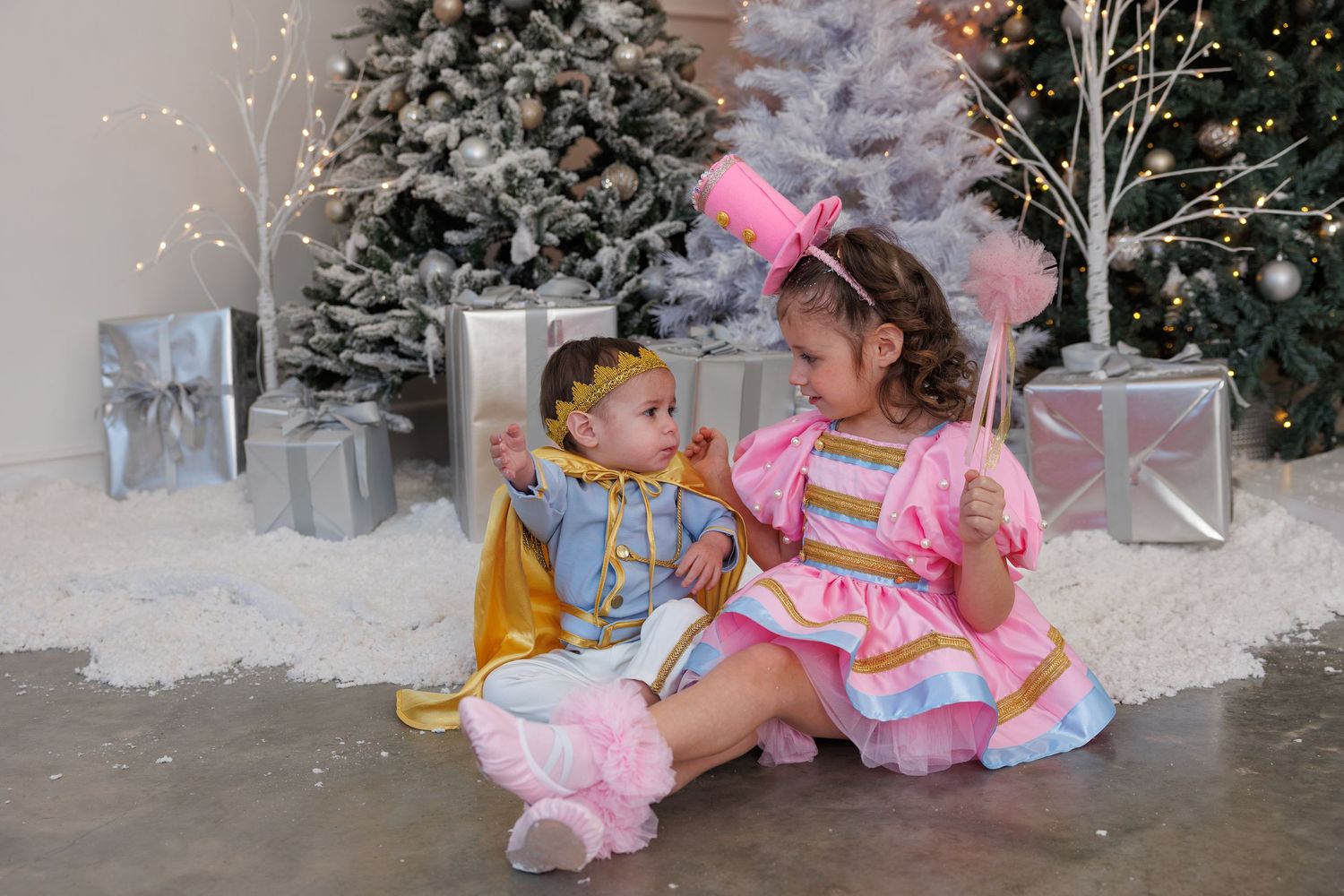 Two children in whimsical costumes sit together among snowy Christmas decorations and silver wrapped presents.