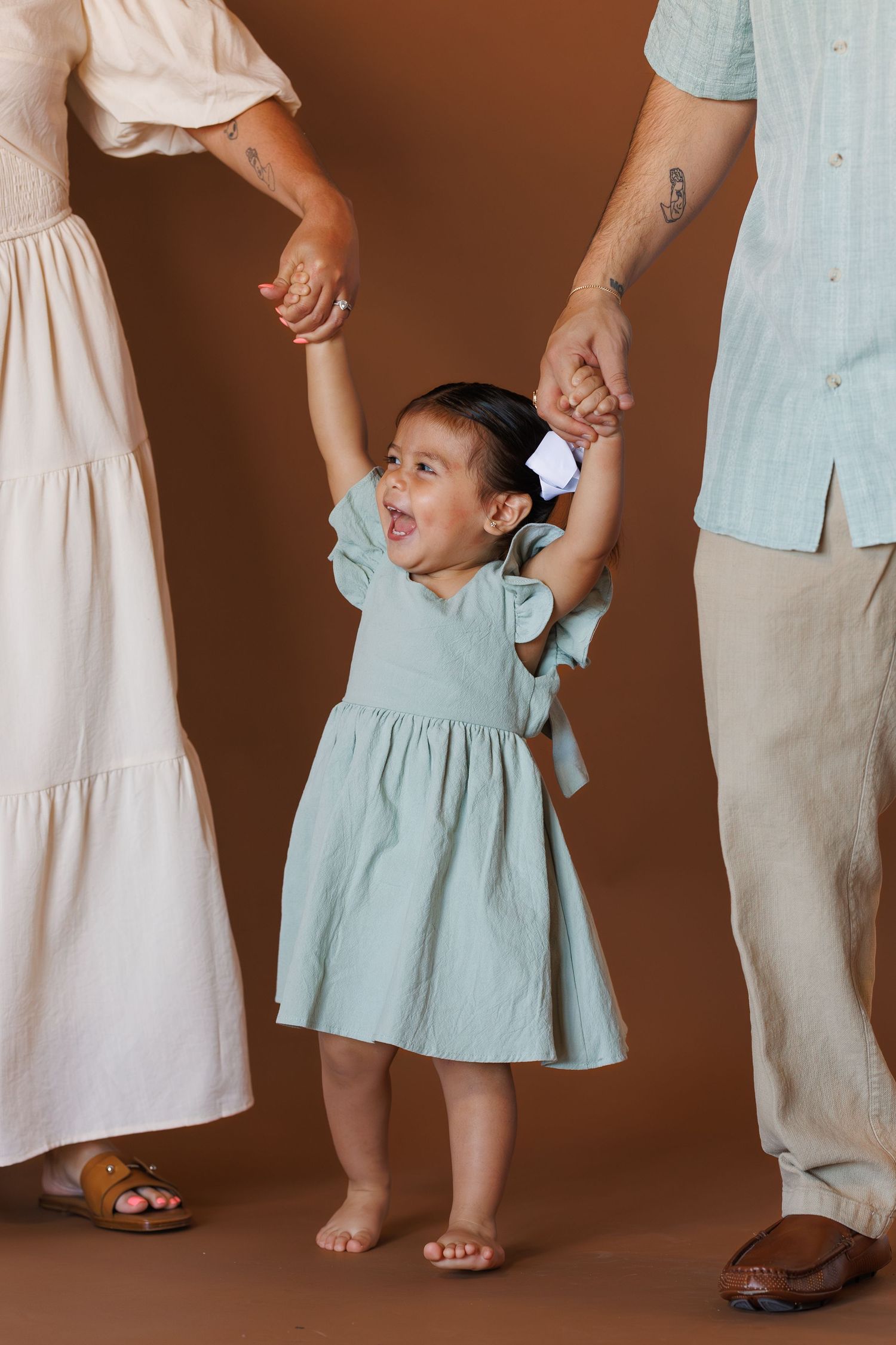Toddler girl in mint dress holds parents' hands, smiling joyfully against a warm brown background.