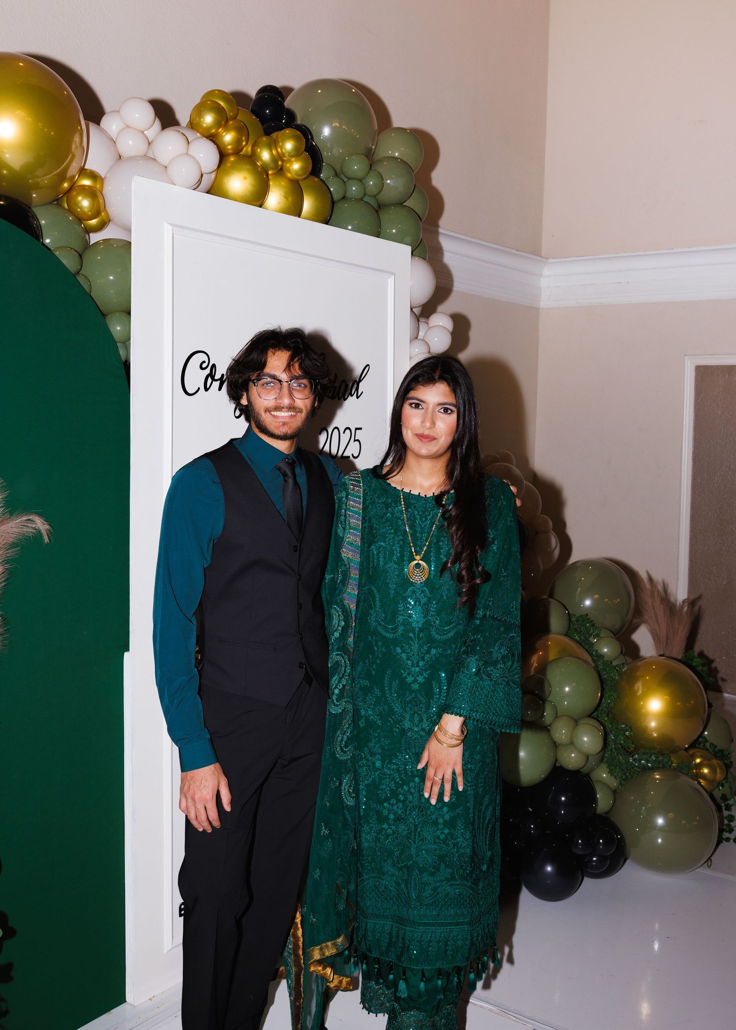 A festive party scene featuring a backdrop of green, gold and white balloons with people posing for photos.