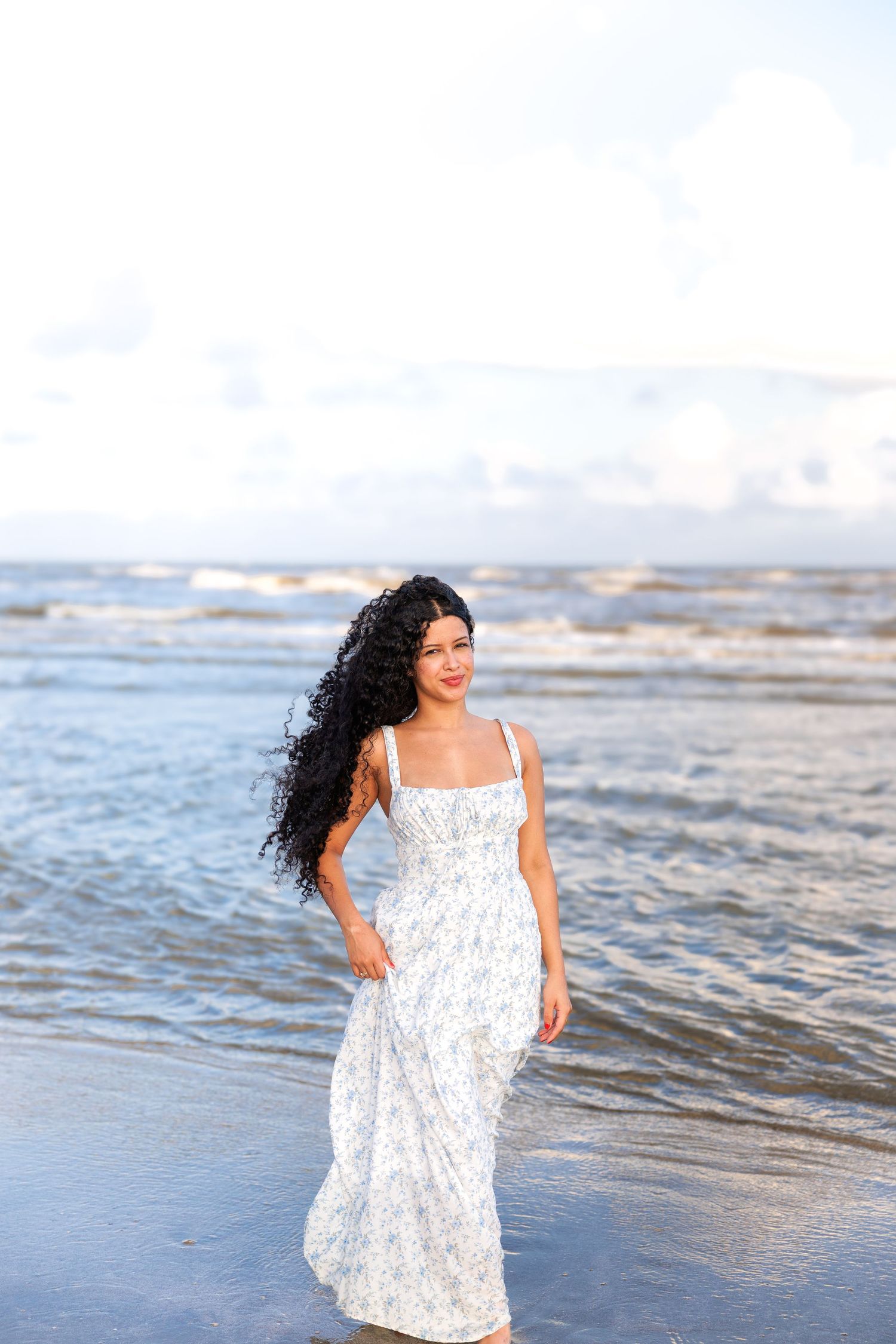 A person in a white dress walks along the shore as waves break on the beach.