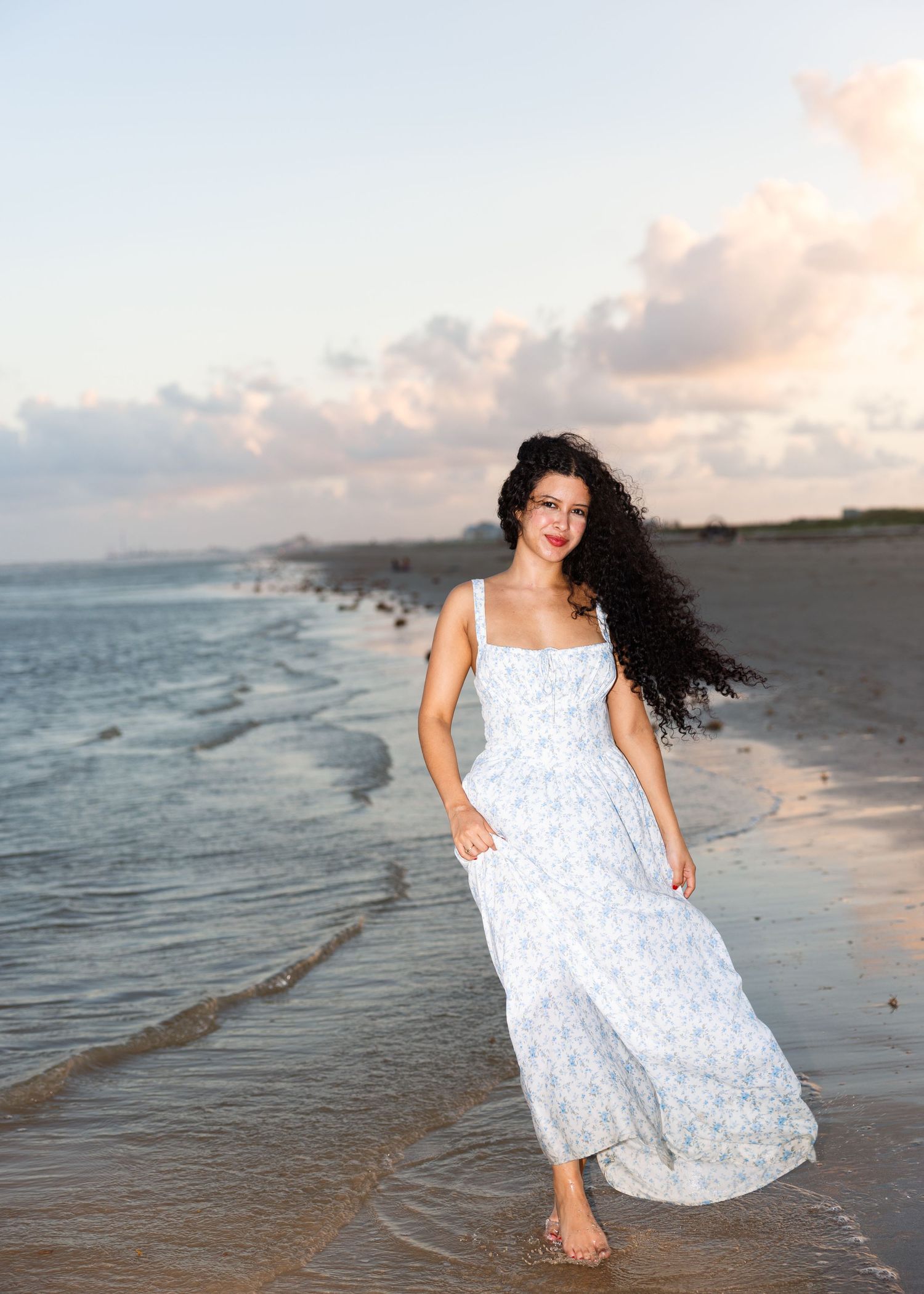 A person in a flowing white maxi dress walks barefoot along a scenic beach shoreline at sunset.
