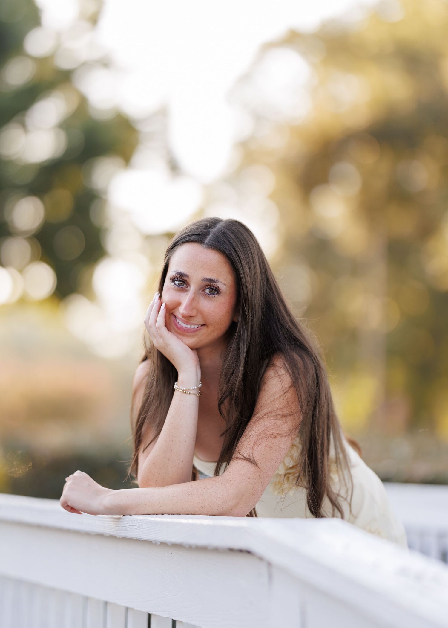 Smiling young woman with long brown hair rests her chin on her hand while leaning on a white railing in warm golden-hour light.