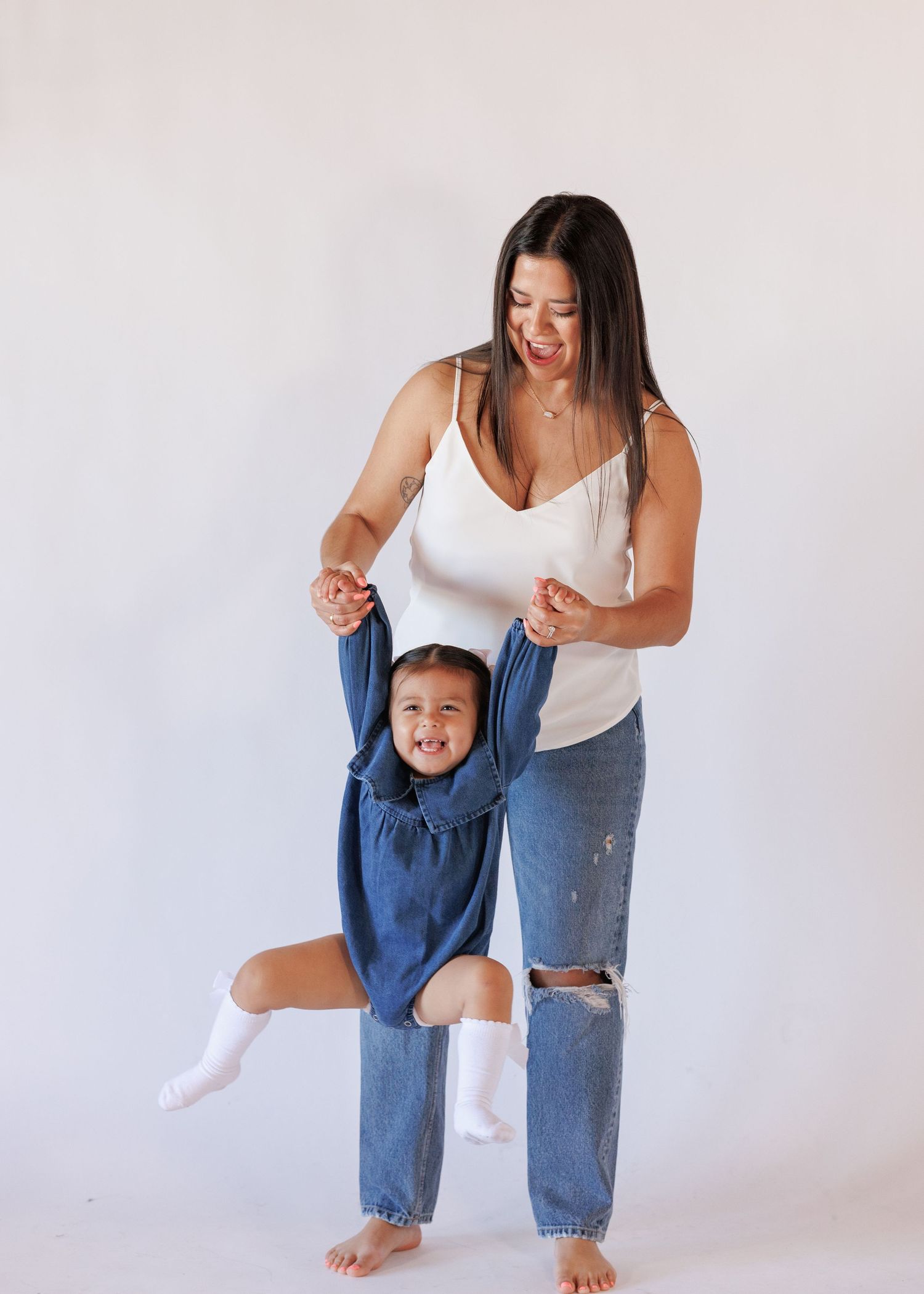 A woman in white top and jeans playfully lifts a smiling toddler in a blue dress against a white background.