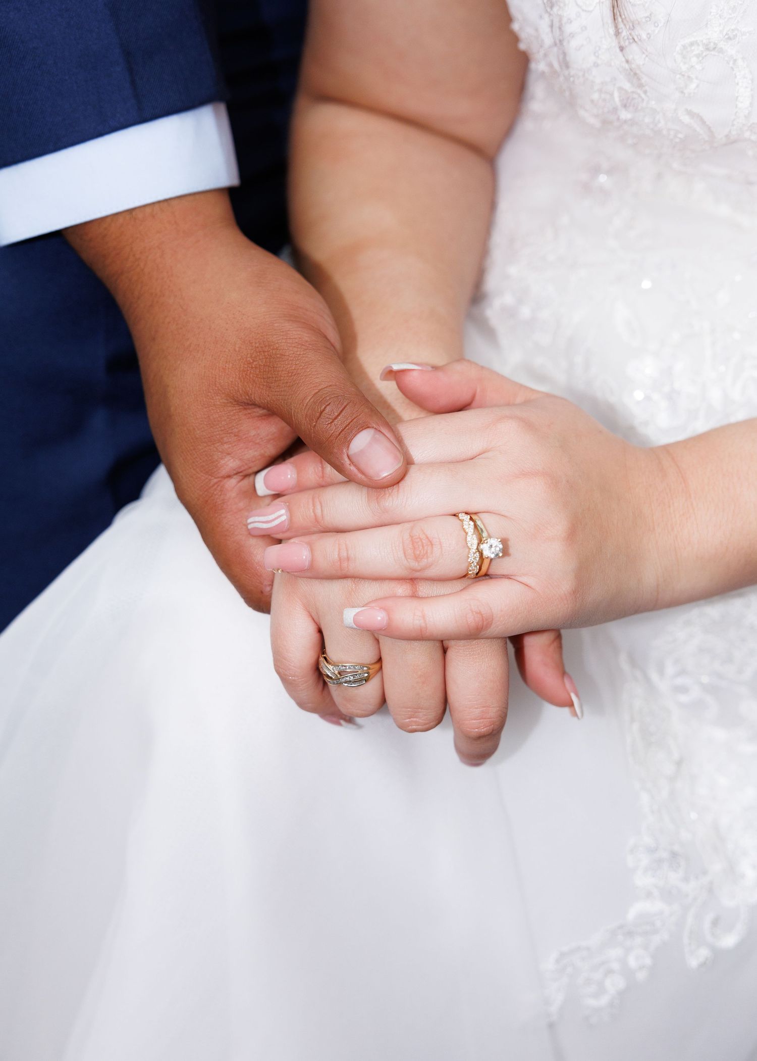 A close-up of newlyweds' hands with wedding rings, showing interlocked fingers against white lace dress and navy suit.