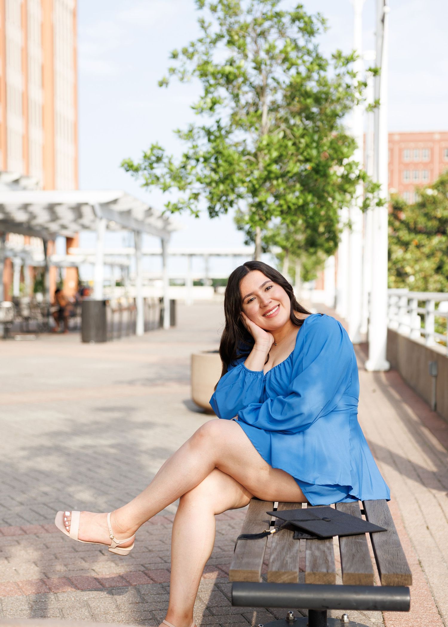 Someone in a blue dress sitting casually on a bench along a brick walkway on a sunny day.