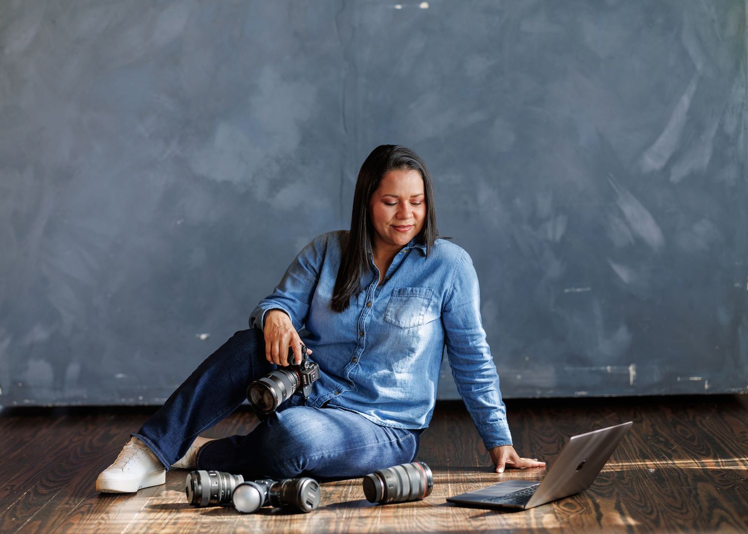 Woman in denim outfit sitting on floor with camera and laptop, reviewing photos in a modern studio.