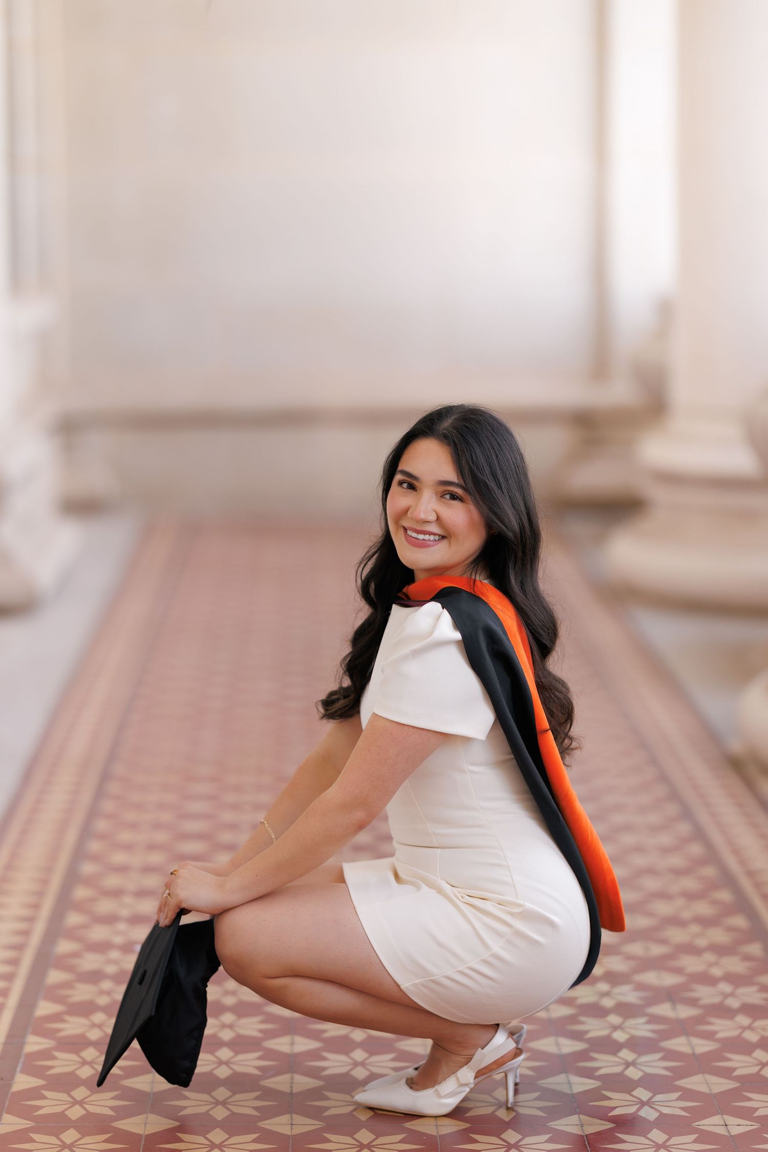 Smiling young woman in white dress and graduation regalia crouching on ornate tile floor in elegant columned hallway.