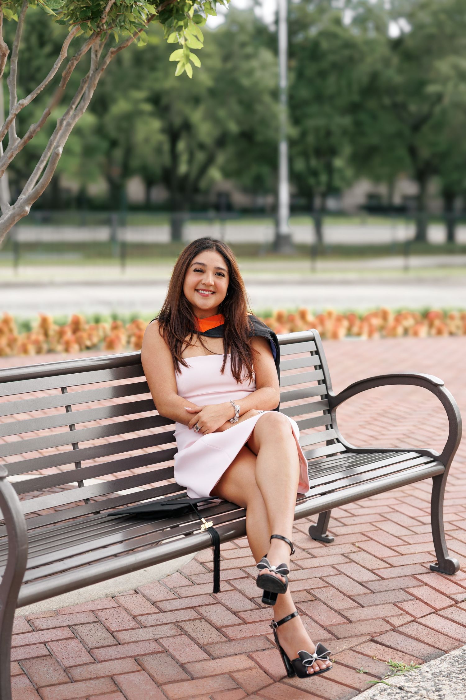 Young woman in pink dress and orange scarf sits smiling on a park bench, with green trees and flowers in background.