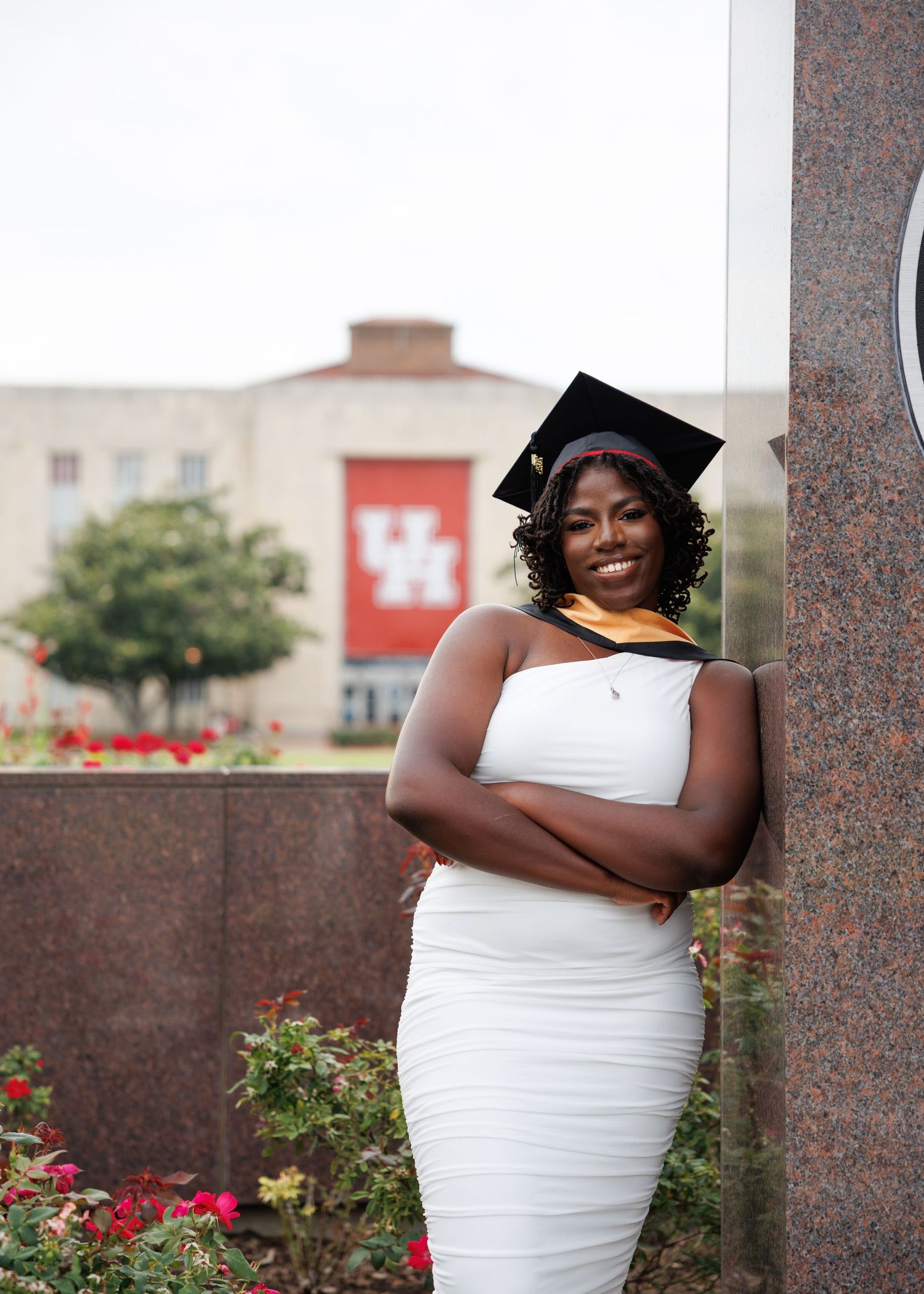 Graduate in white dress stands near stone column with red flowers and university building in background.