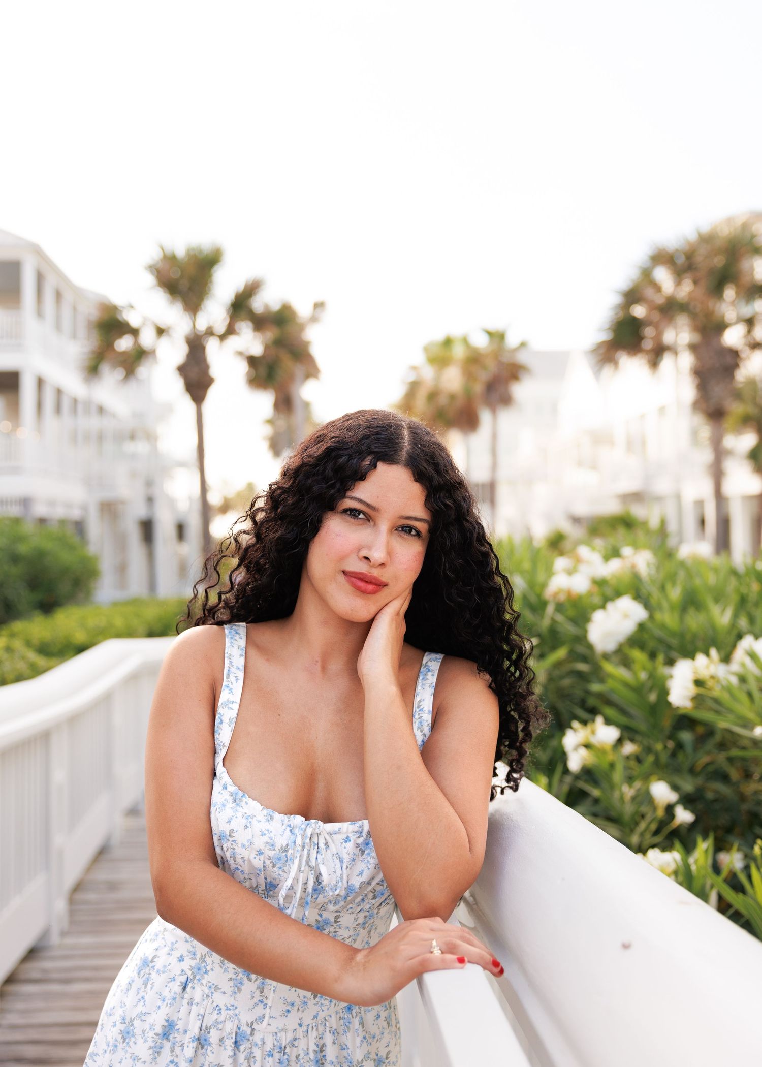 A person in a floral summer dress poses on a white boardwalk with palm trees and white buildings in the background.