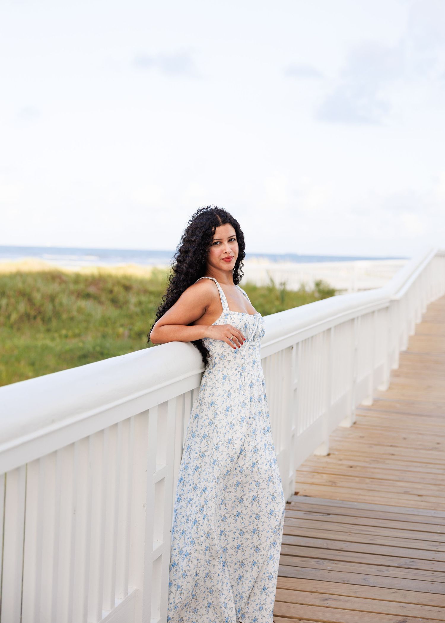 Someone in a flowy floral maxi dress poses on a wooden boardwalk railing near a coastal beach landscape.