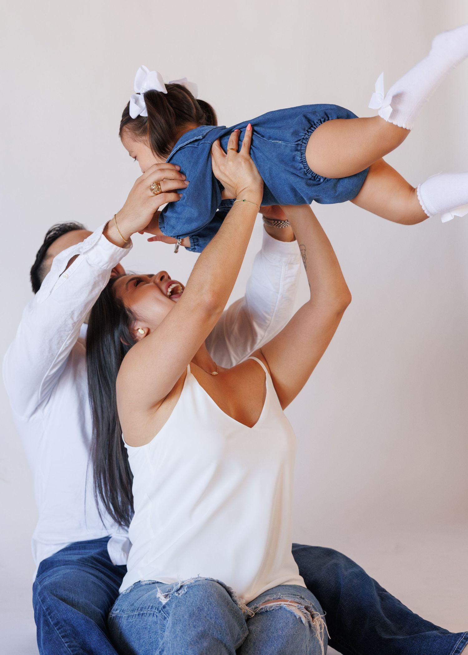 Happy family with parents in white tops and jeans lifting their toddler girl in denim dress with white bow.