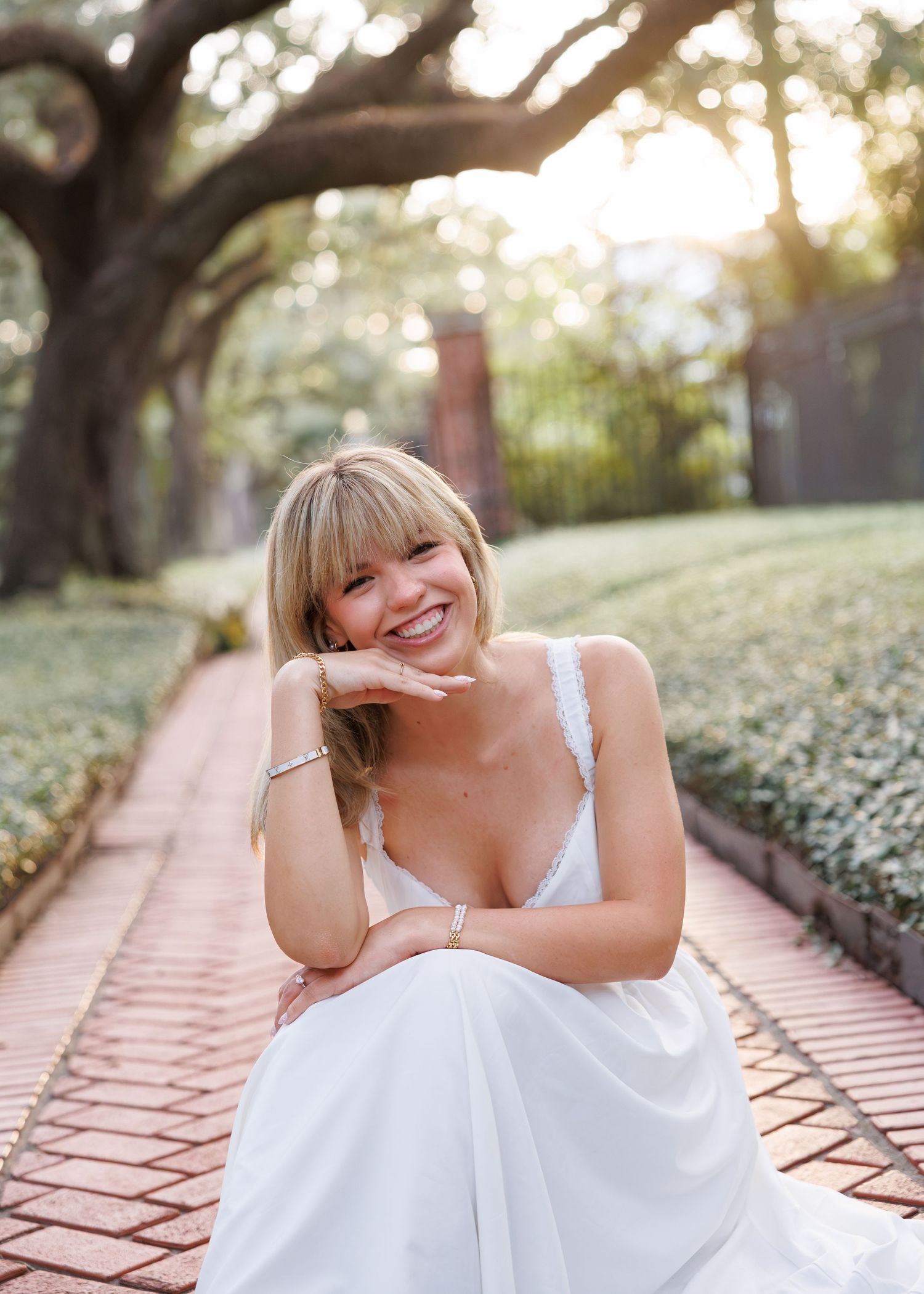 Smiling woman with blonde bangs in a white sundress sits on a brick pathway lined with greenery under sunlit oak trees.