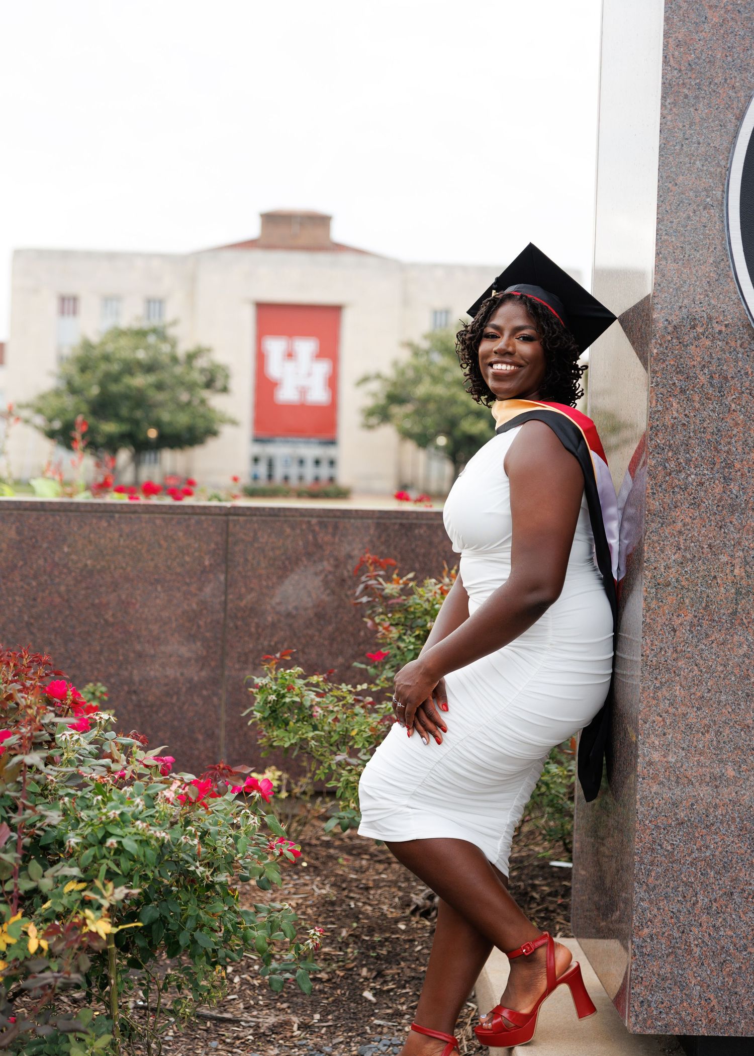 Graduate poses in white dress and red heels in front of University of Houston building with red banner and roses.
