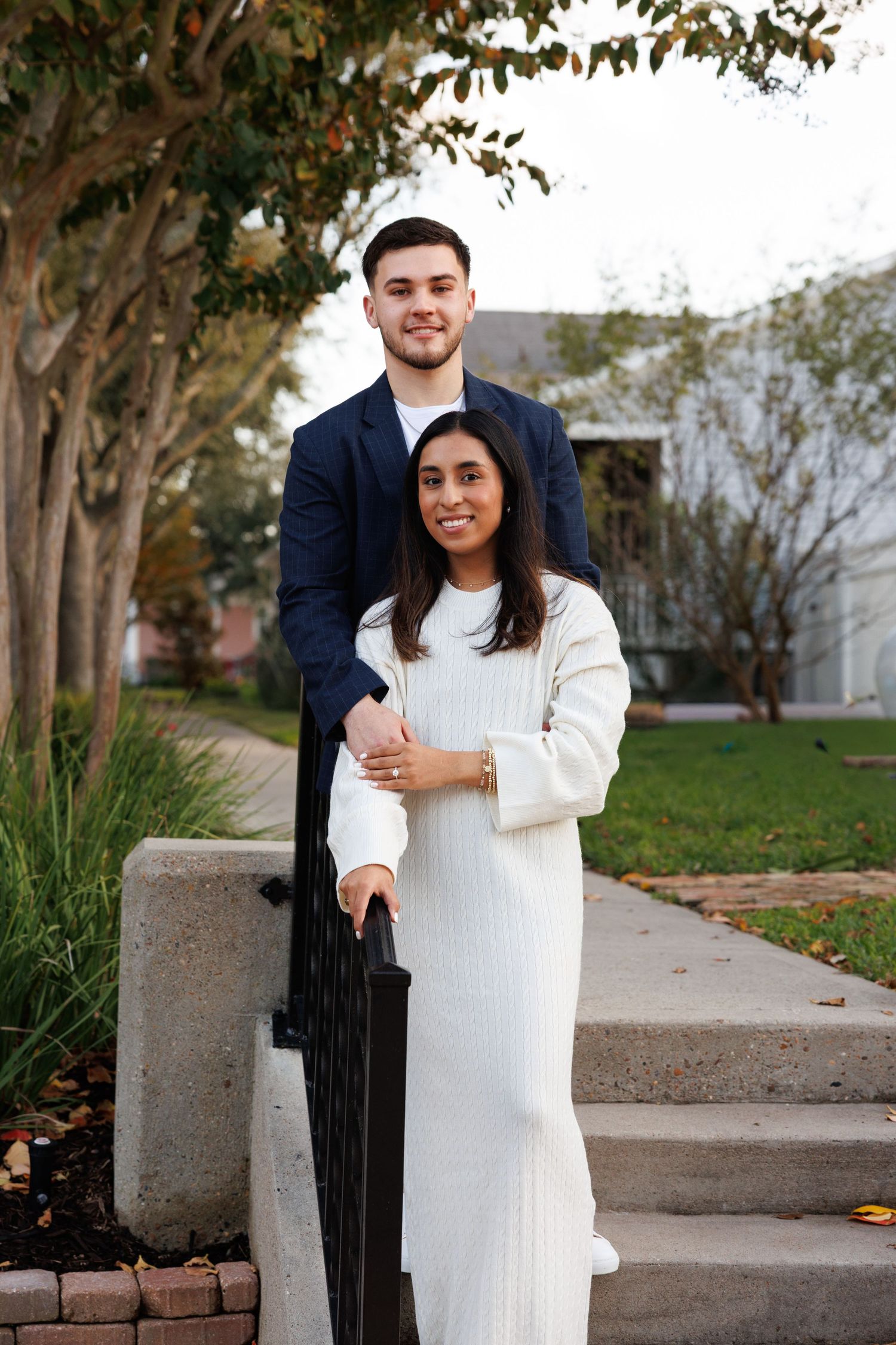A couple poses together outdoors on a fall day with trees and brick landscaping in the background.