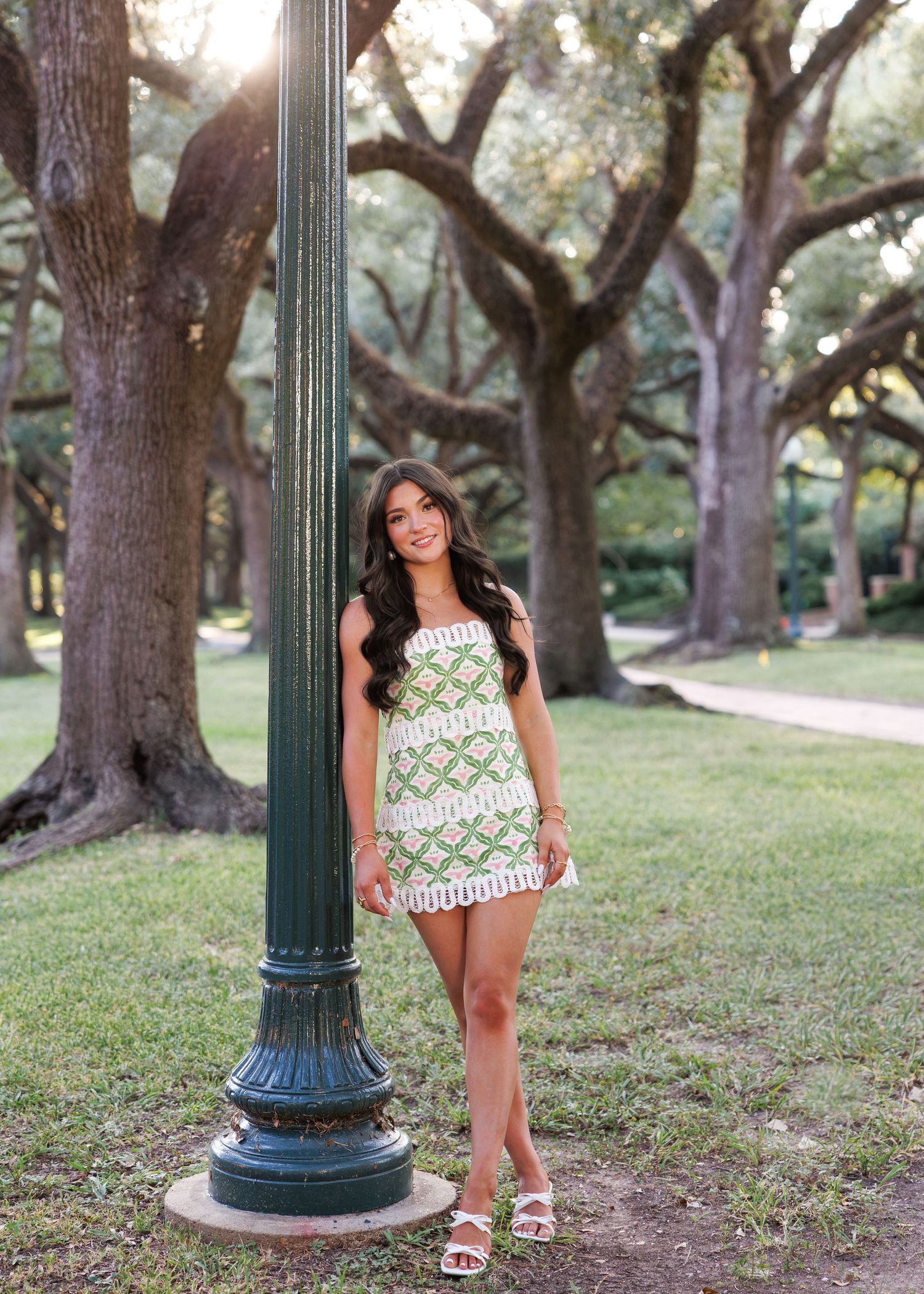 Young woman in green patterned romper posing by a lamp post in a lush park with large oak trees.