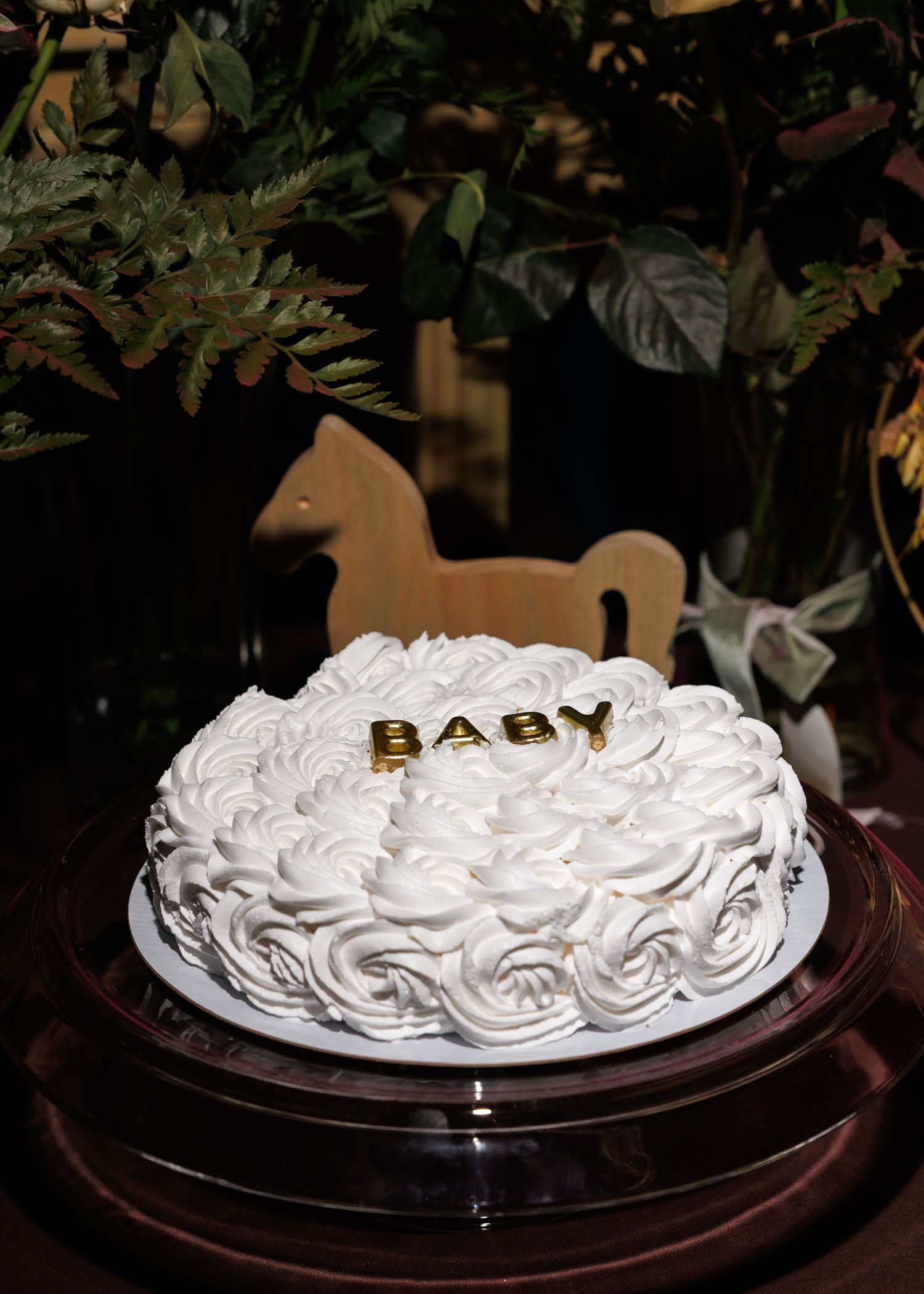White frosted cake with rosette decorations and gold 'BABY' text on top sits on dark wooden stand with greenery backdrop.