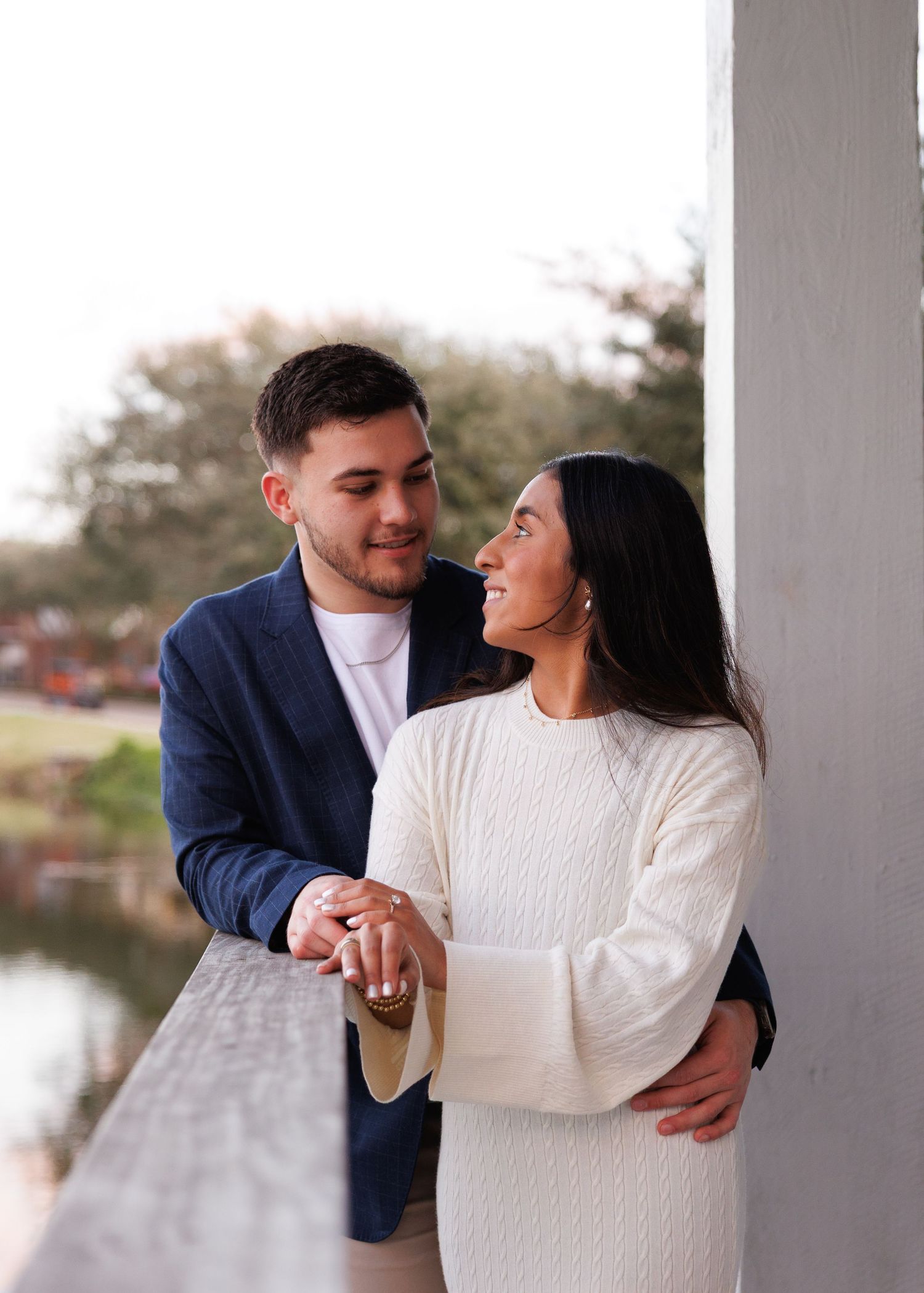 A couple in coordinated white and navy outfits shares romantic moments during their engagement photoshoot by a lake.
