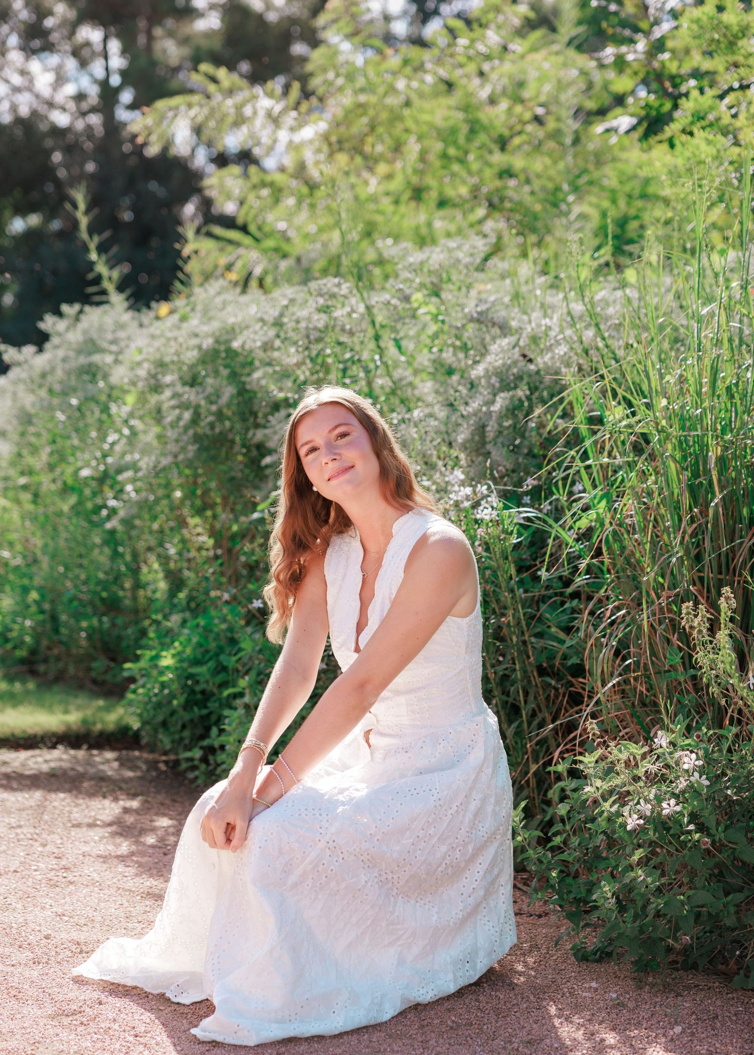 Smiling woman with long hair sits on a gravel path in a white sleeveless dress, surrounded by lush green garden foliage in sunlight.