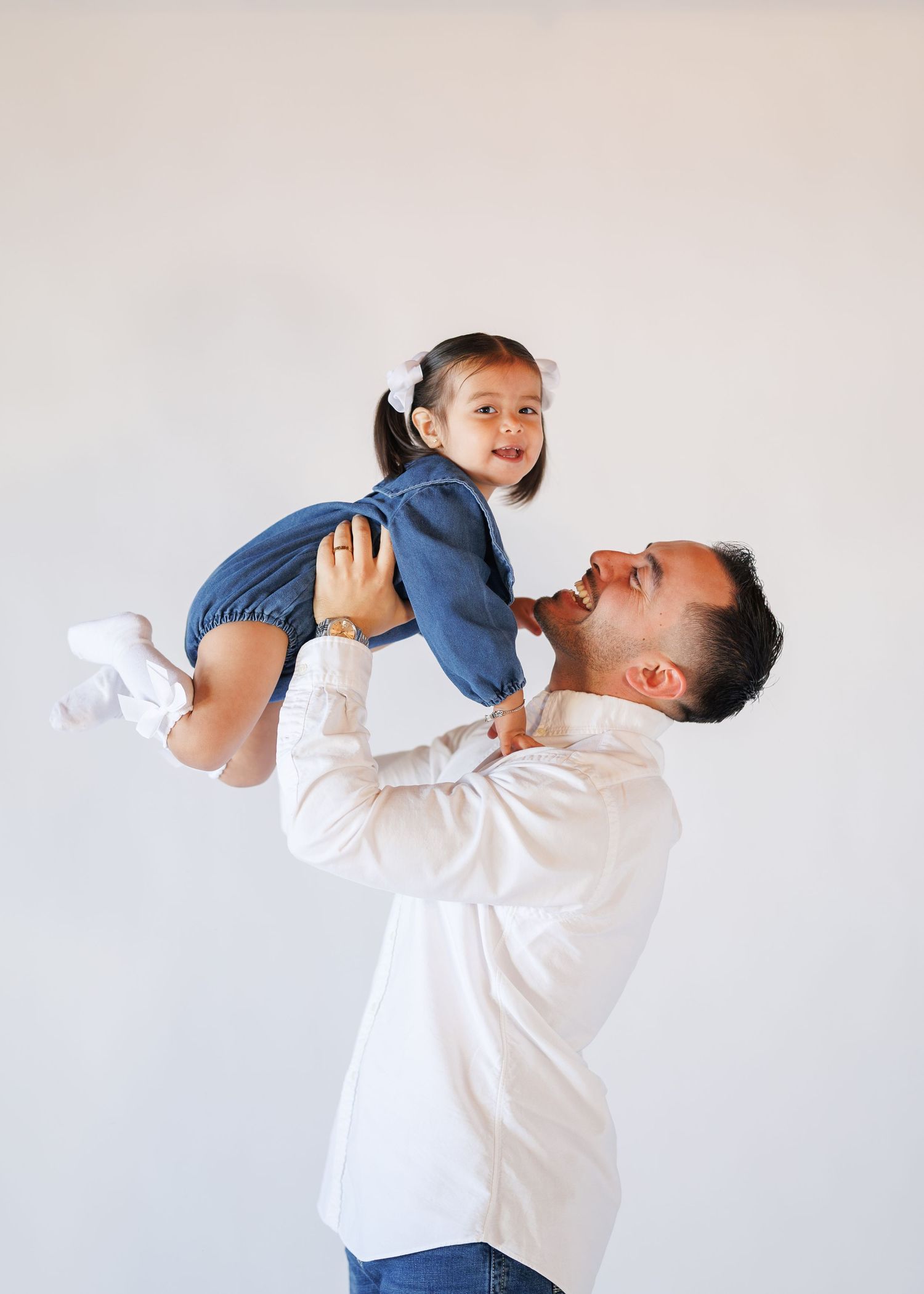 A father joyfully lifts his smiling toddler daughter in the air against a white background.