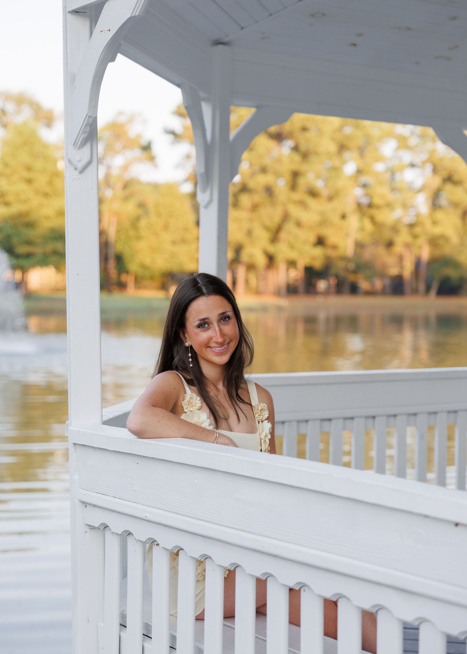 Smiling young woman with long dark hair leans on a white gazebo railing beside a calm lake with golden sunlit trees behind her.