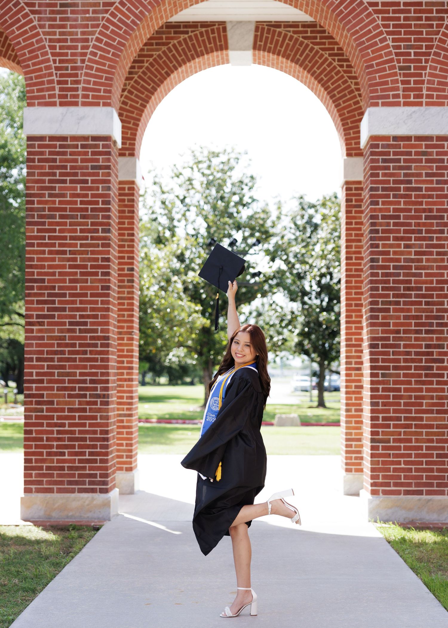 A graduate in cap and gown celebrates under a red brick archway on a university campus during commencement festivities.