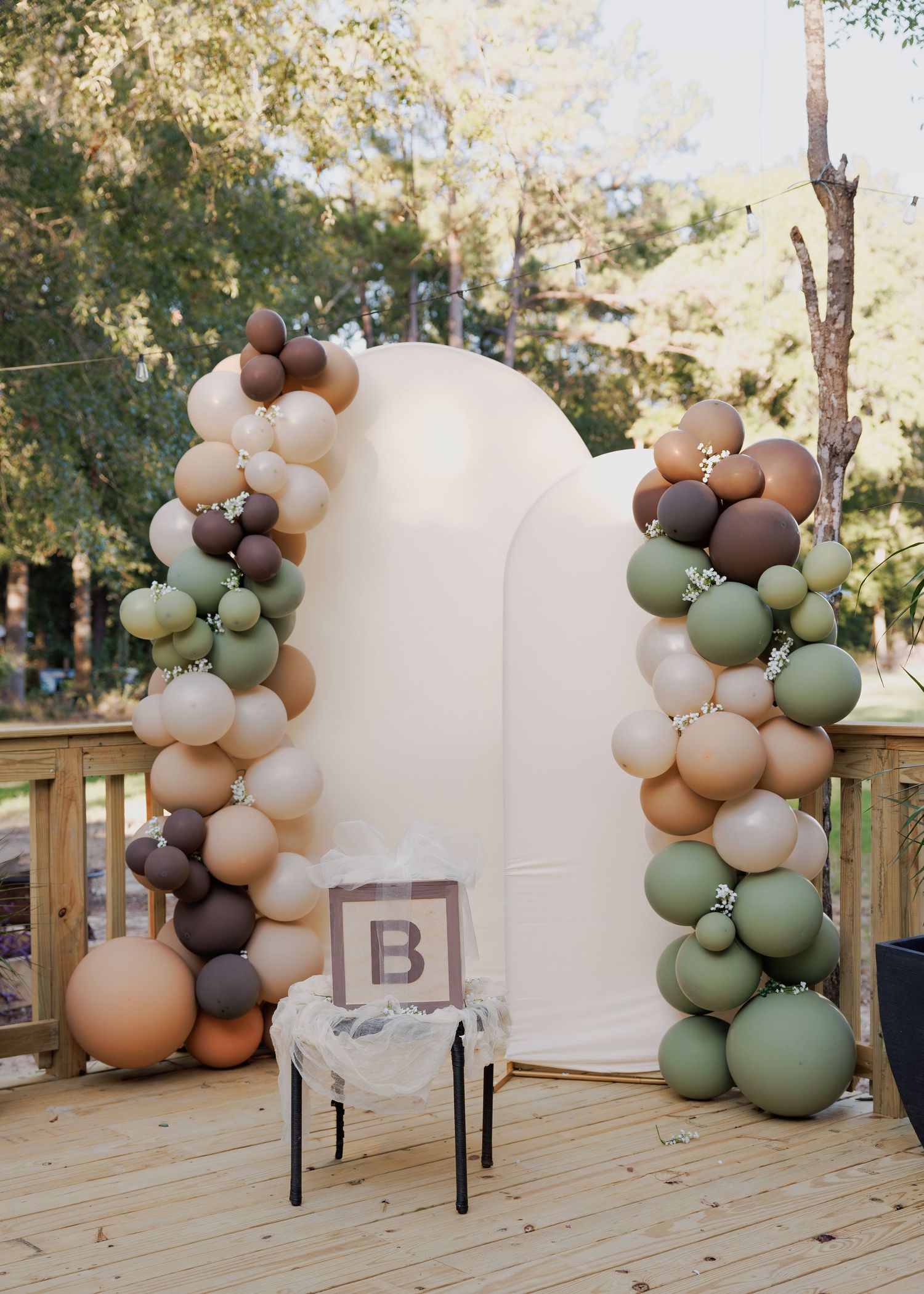 Neutral-toned balloon arch installation with sage green, beige, and white balloons decorating an outdoor wooden deck setting.