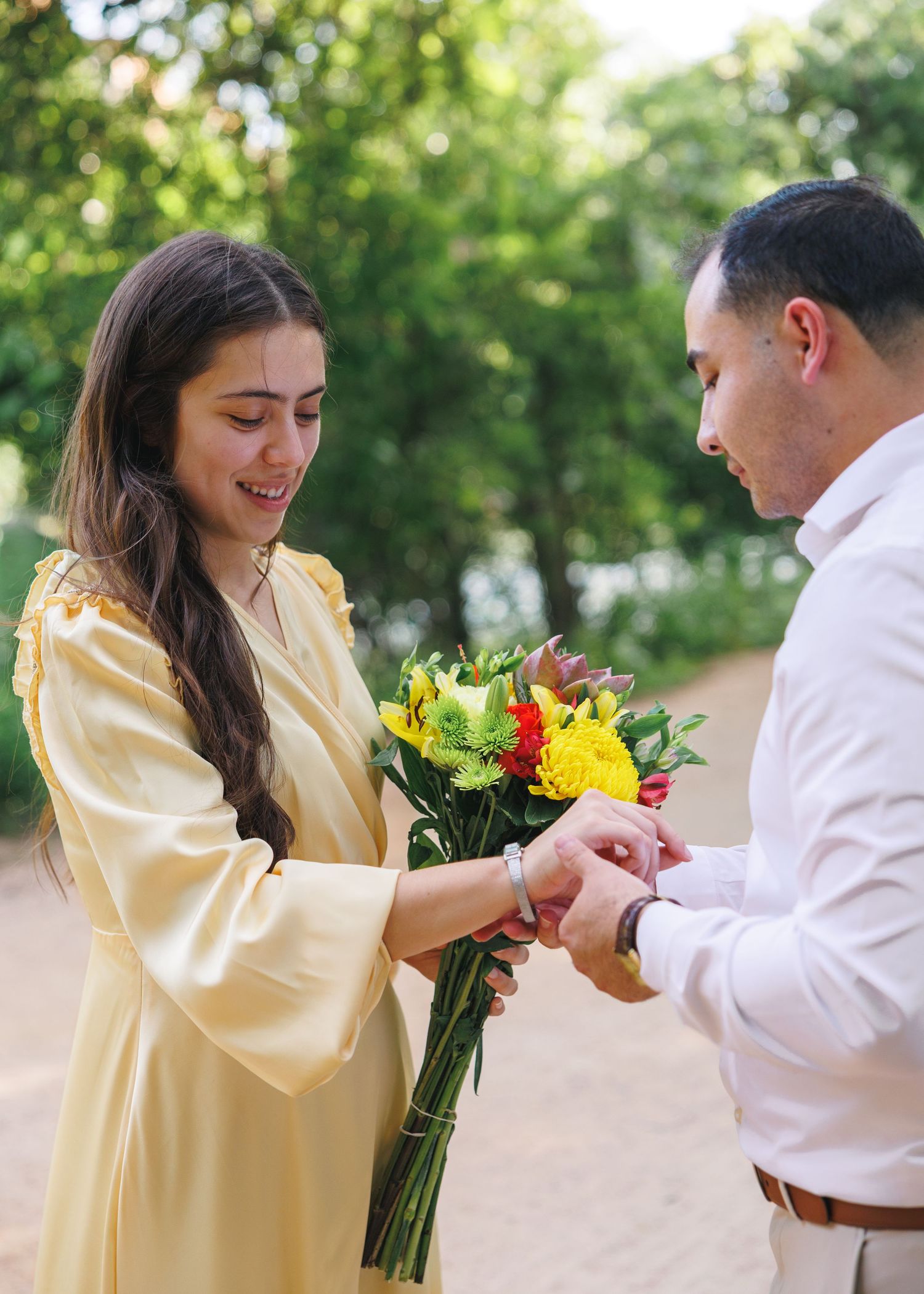 Man placing ring on woman's finger as she holds colorful bouquet in sunny park, romantic proposal moment.