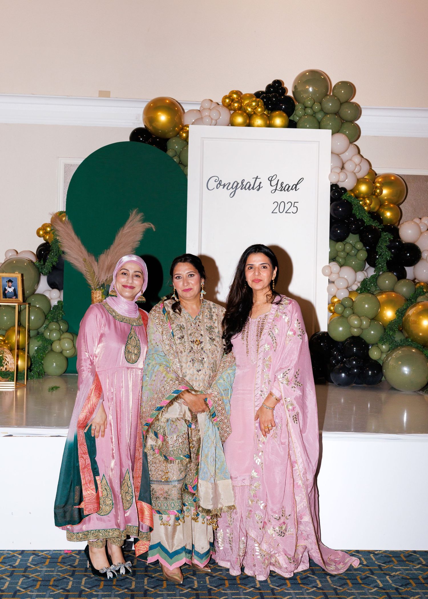 Elegant celebration setup with green and gold balloon arch, decorative backdrop, and attendees in pink and beige formal attire.