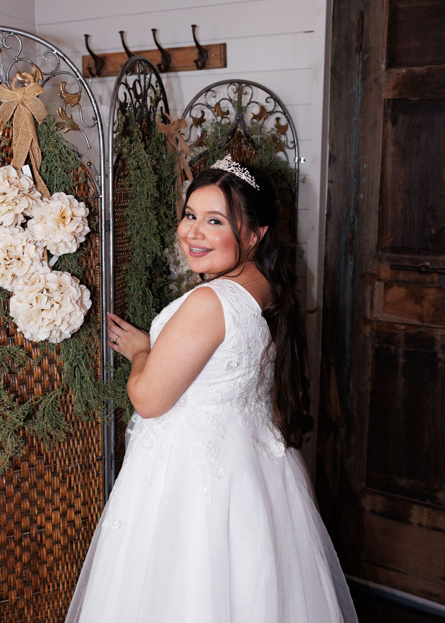 Beautiful bridal gown in white lace and tulle with ornate crown accent standing near decorative wall with floral arrangements.