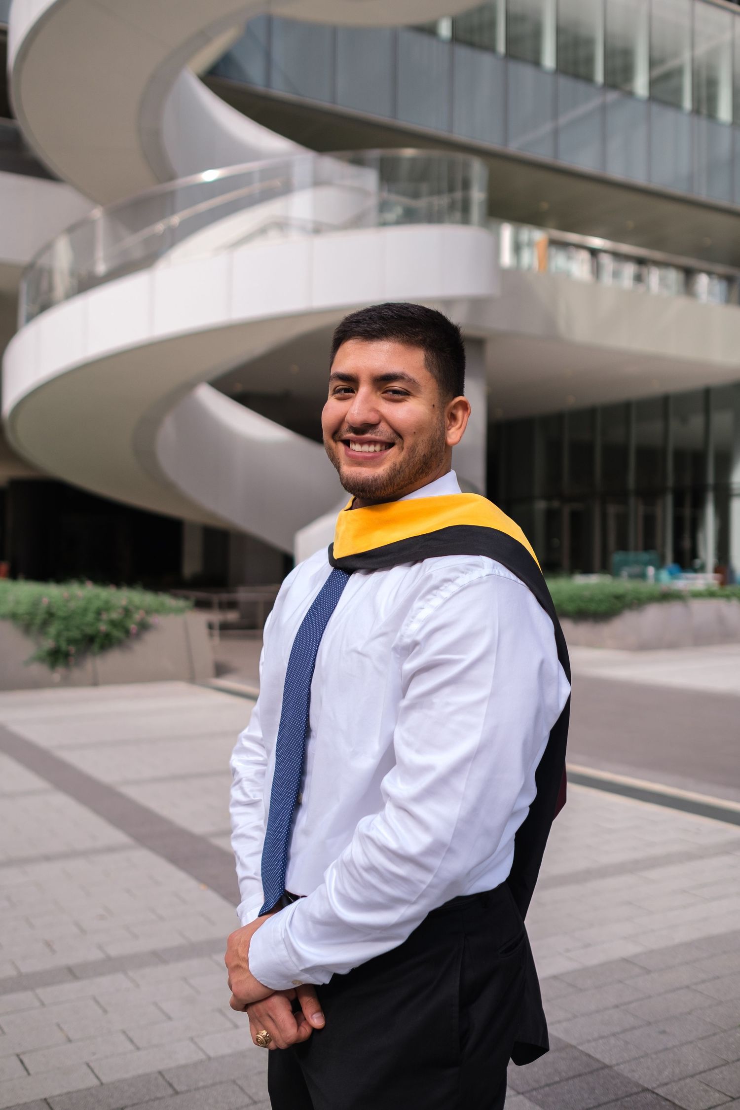 A person wearing a white dress shirt and navy vest smiles while standing near a modern architectural building.