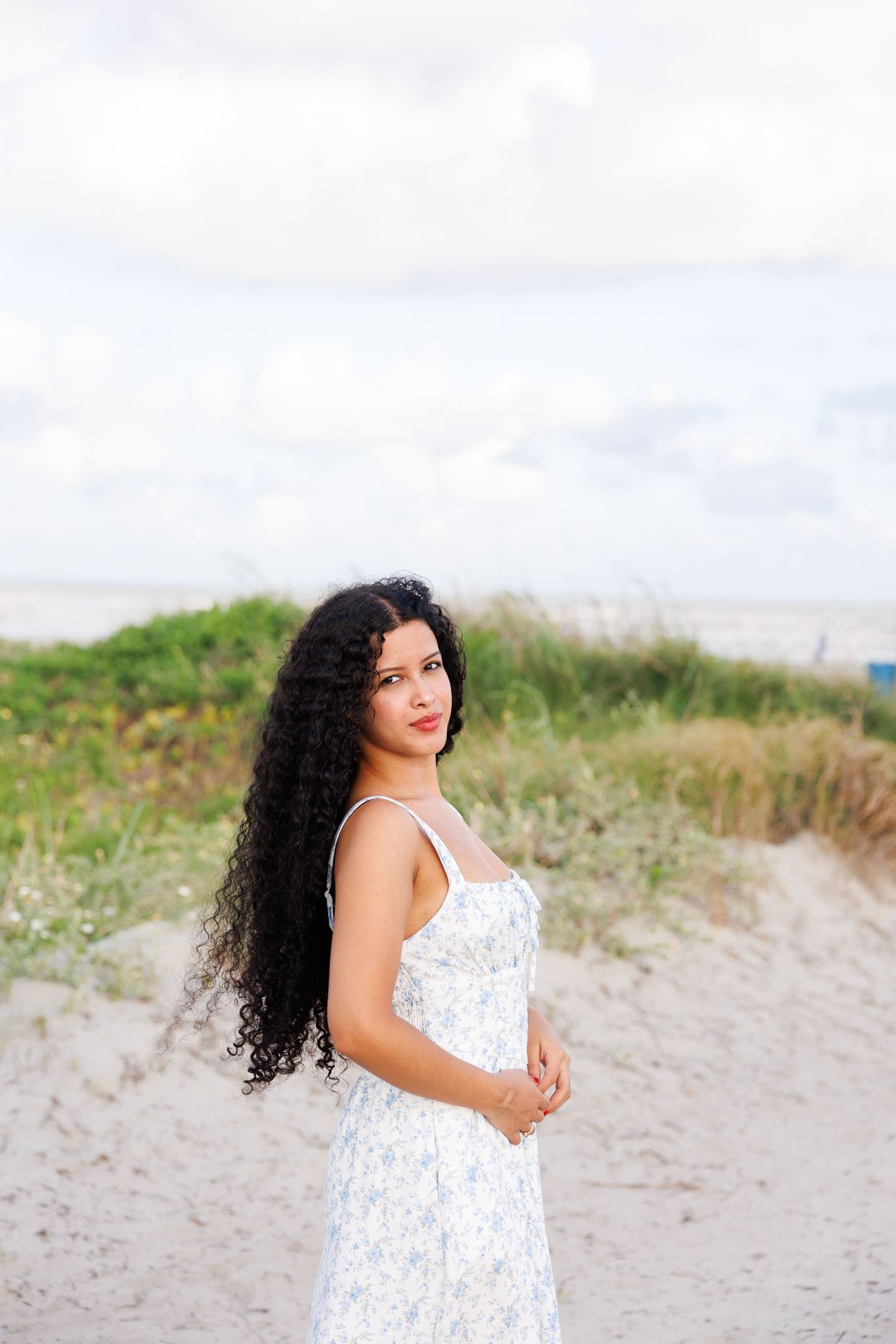 A woman in a floral white summer dress poses gracefully on a sandy beach path surrounded by green grass.