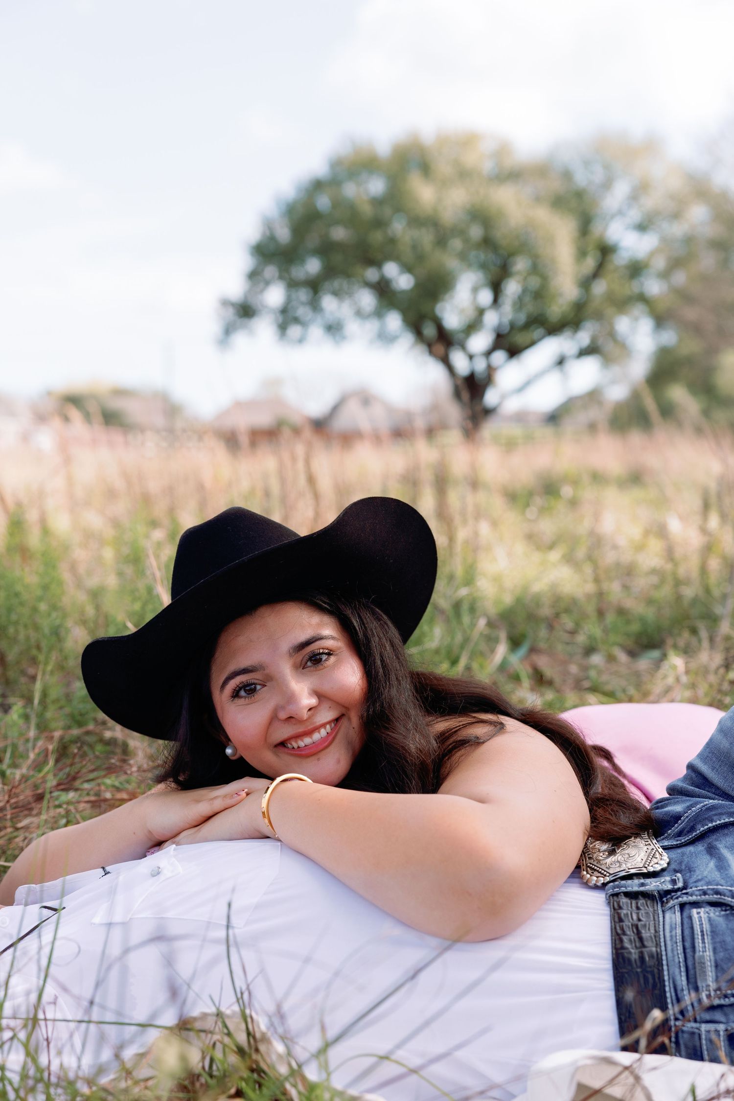 Smiling woman in black cowboy hat lying in a grassy field wearing denim jacket and gold bracelet.