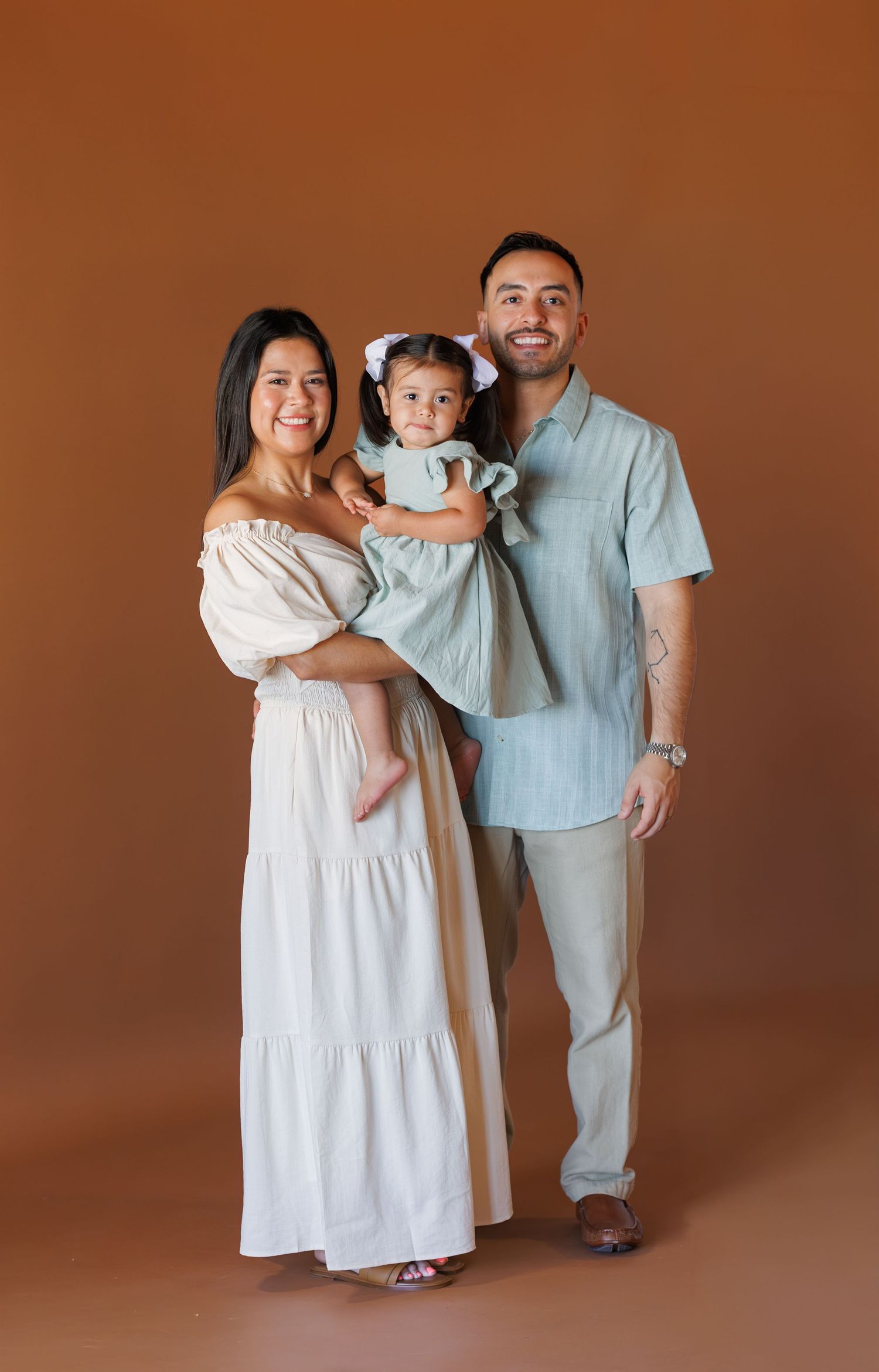 A smiling family of three poses in a studio, with parents holding their toddler daughter against a warm brown background.