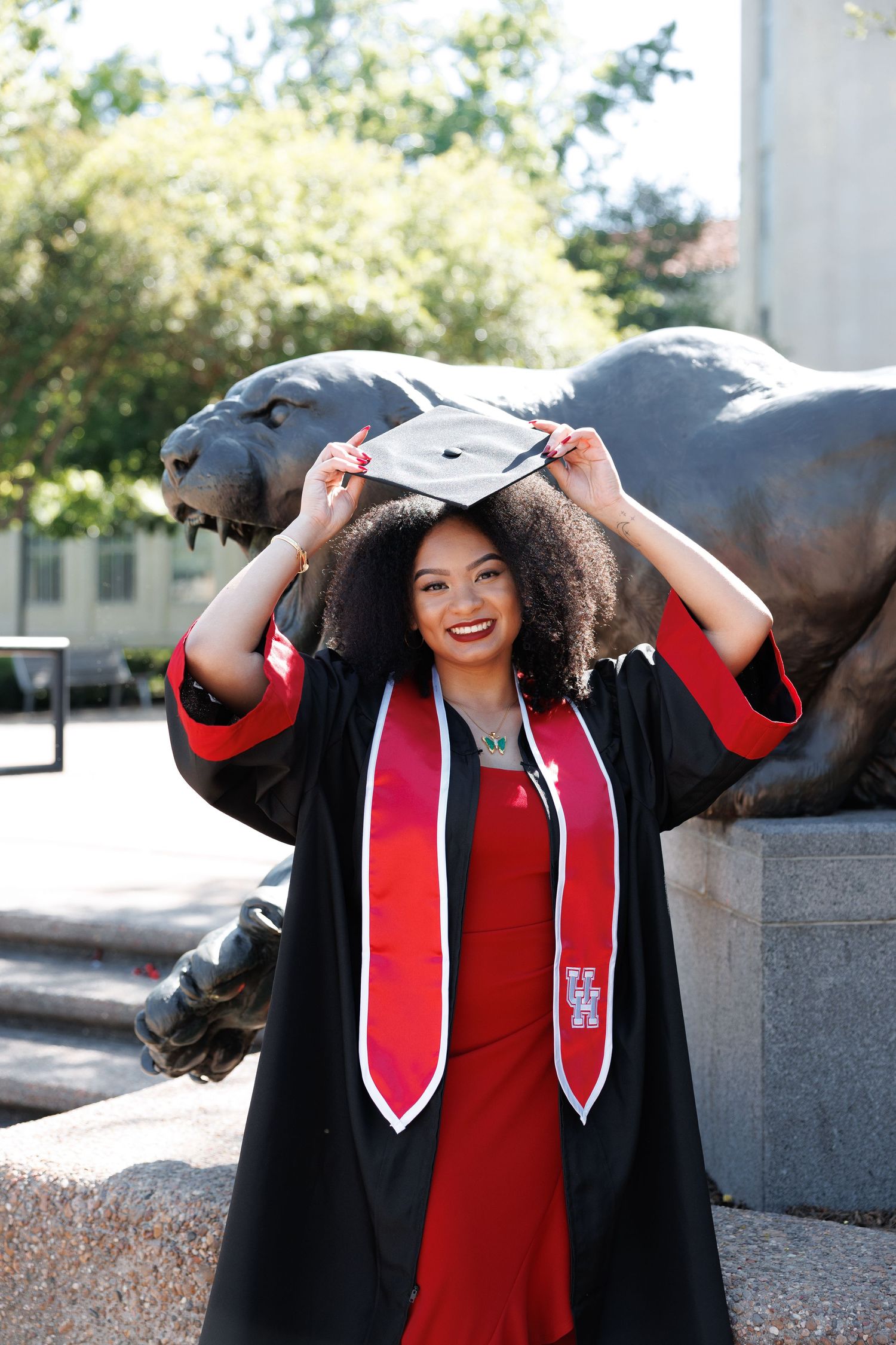 Graduate in a black cap and gown with red stole poses with campus statue during outdoor commencement photoshoot.