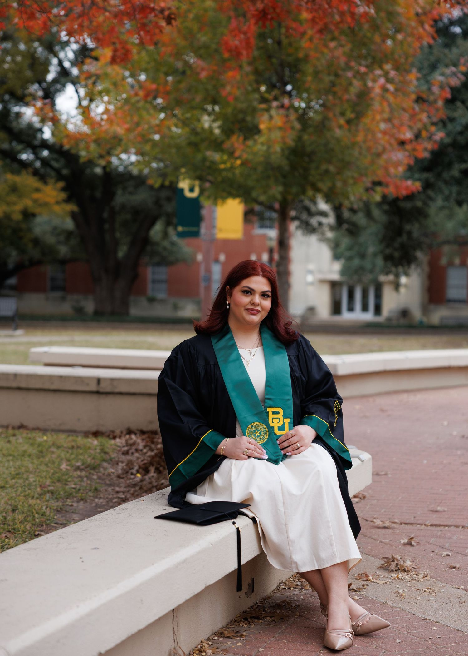 A woman in a green blazer and white dress sits on a concrete ledge surrounded by autumn trees with orange foliage.