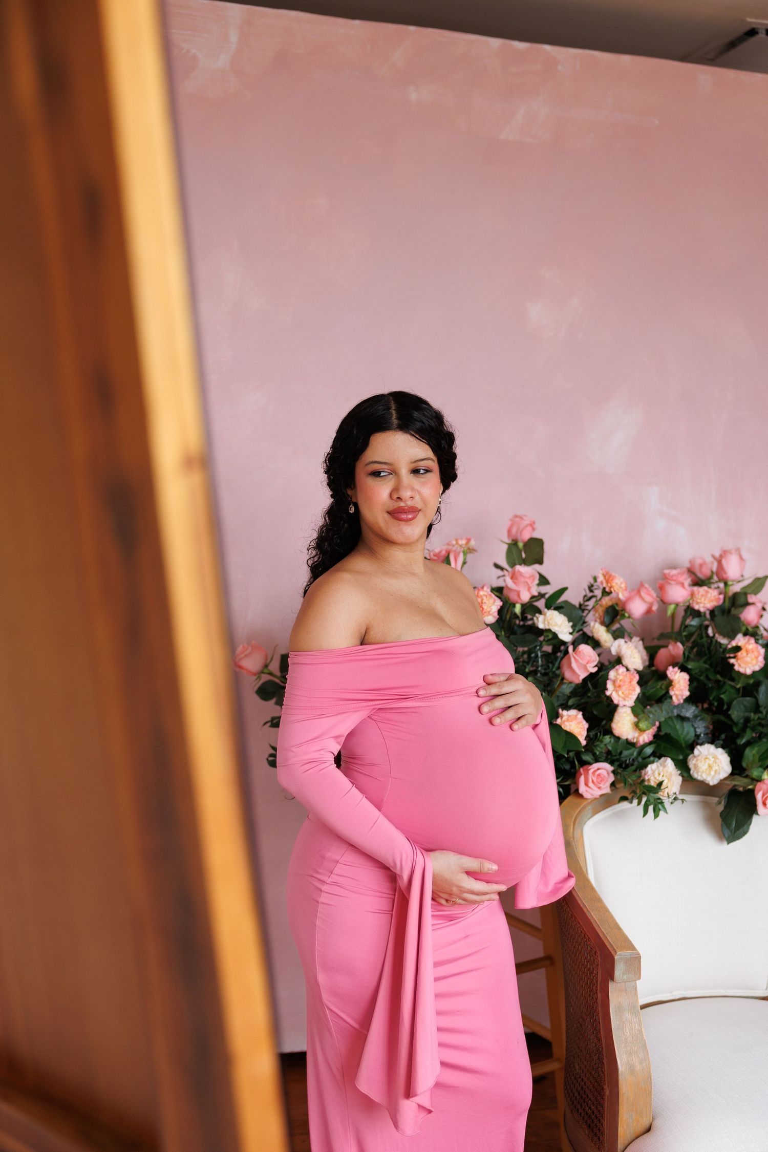 Pregnant woman in pink off-shoulder gown poses gracefully in front of pink roses and matching wall.