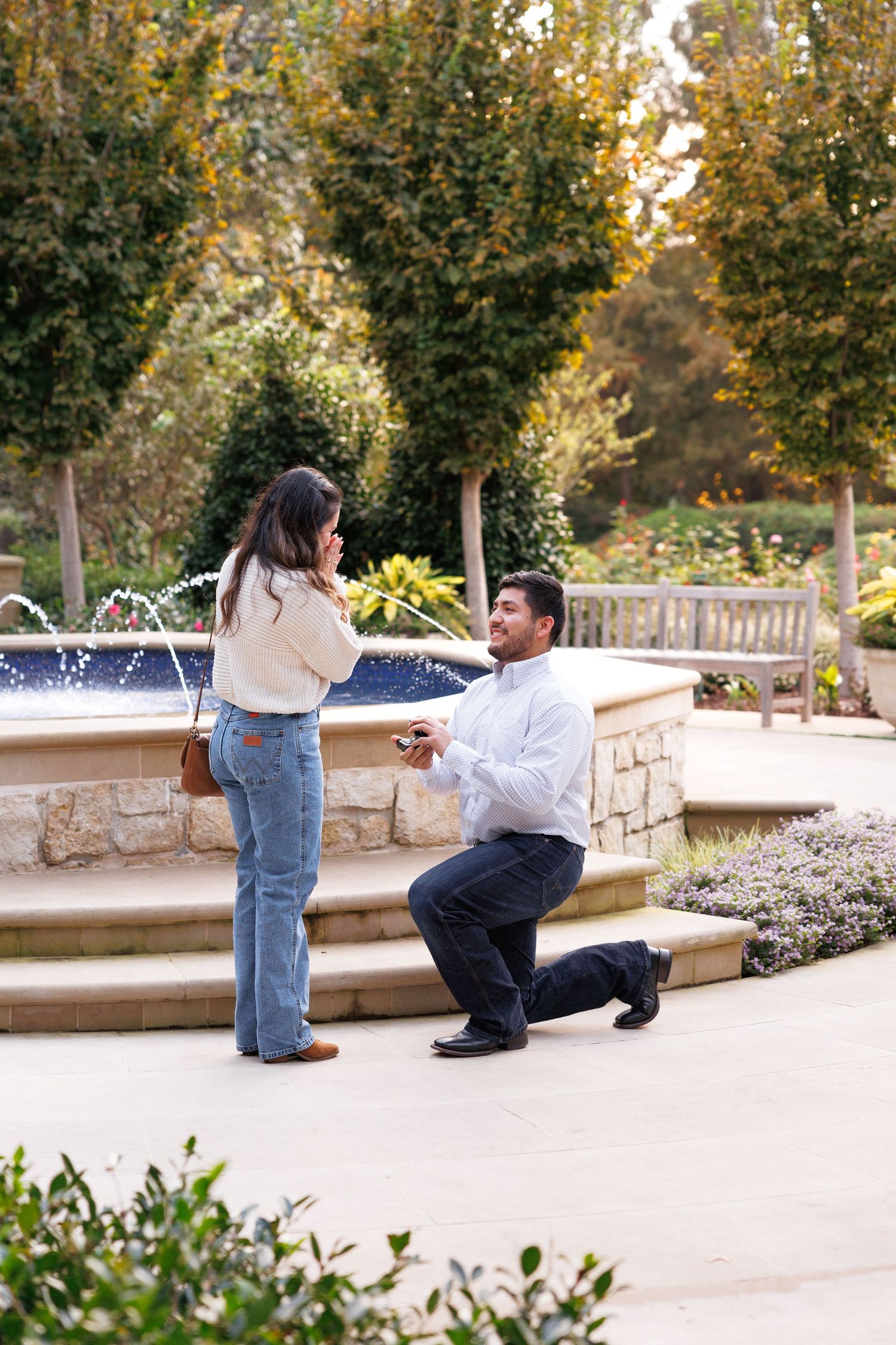 A romantic proposal scene unfolds by a fountain in a garden setting with autumn trees in the background.