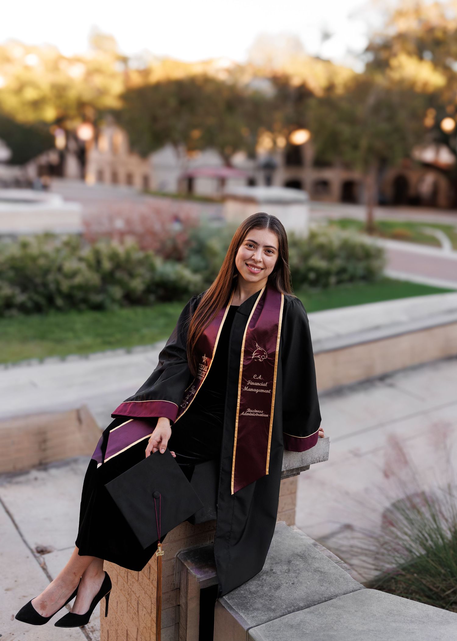 Graduate in black academic regalia with burgundy stole sits on stone ledge during golden hour on university campus.