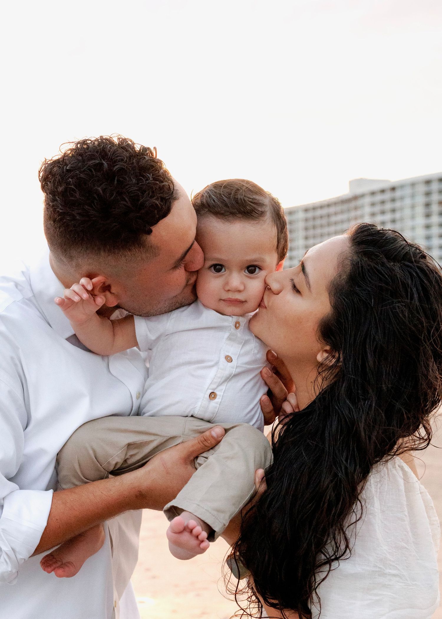 A family shares tender moments of affection and kisses while wearing white clothing against a light background.