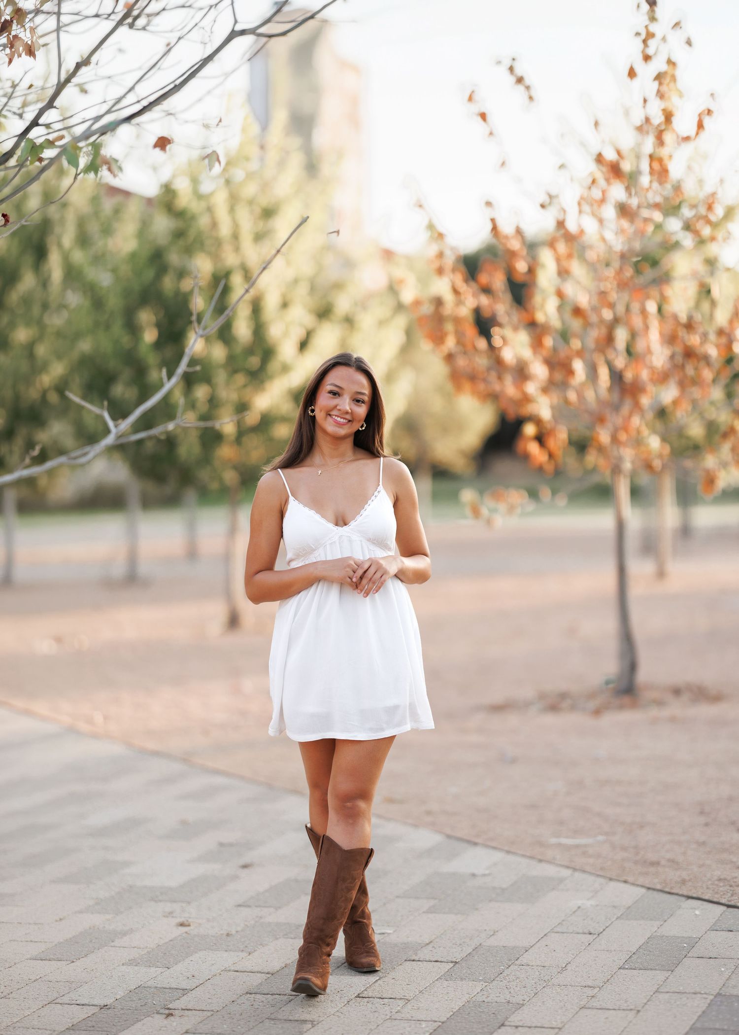 Smiling woman in a white sundress and brown cowboy boots poses on a brick path in a park with autumn trees.