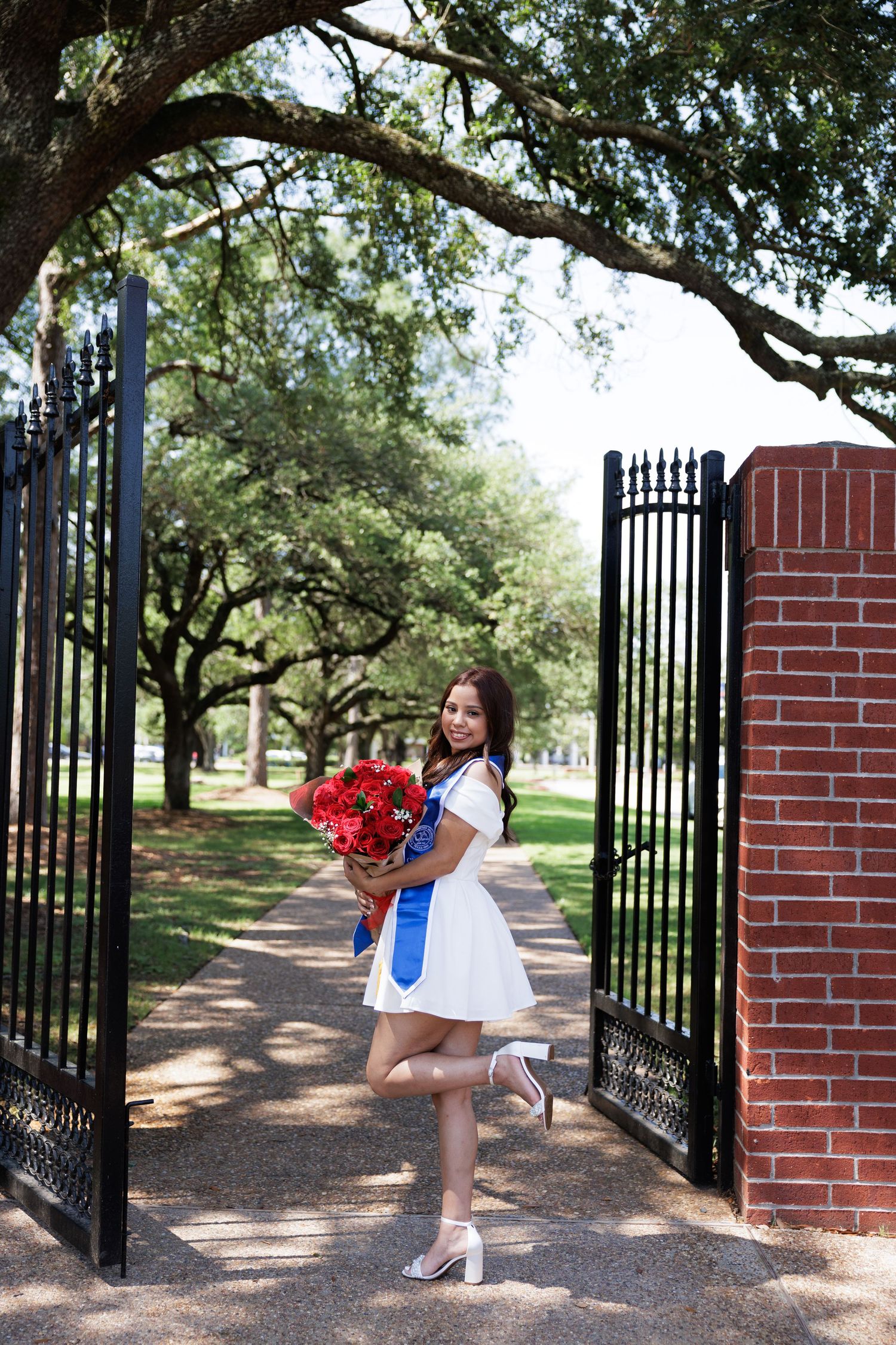 Standing at ornate black metal gates beside brick pillars with red roses and a white dress against a tree-lined path background.