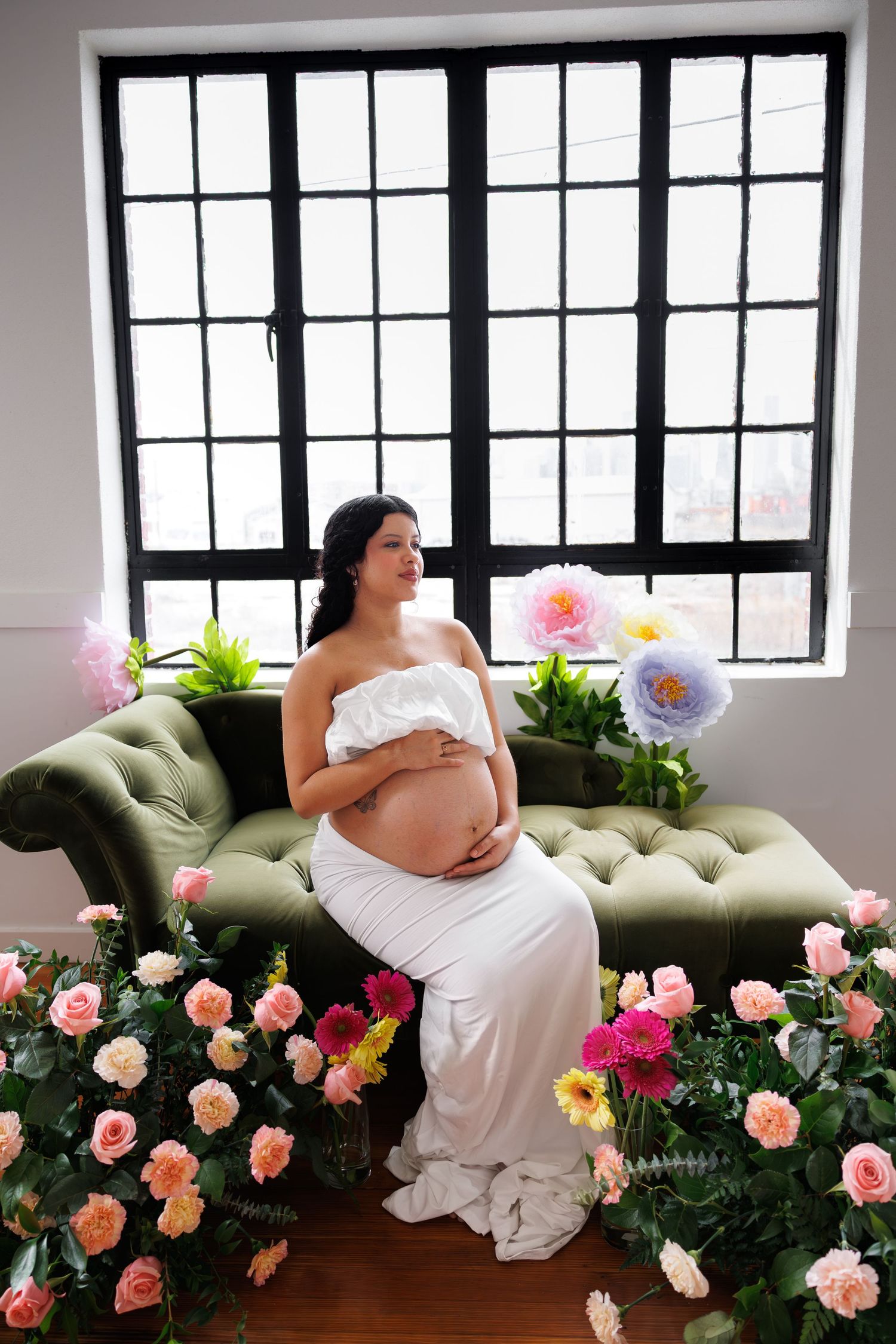 Pregnant woman in white off-shoulder gown sits on green velvet chaise lounge surrounded by colorful roses near large window.
