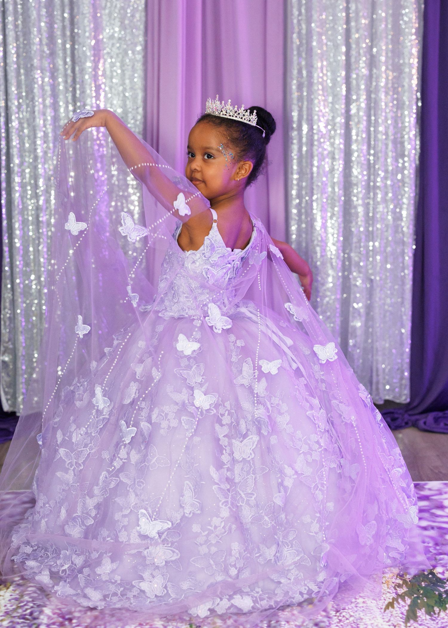 A child in a lavender butterfly ball gown with a tiara strikes a graceful dance pose against a sparkly backdrop.
