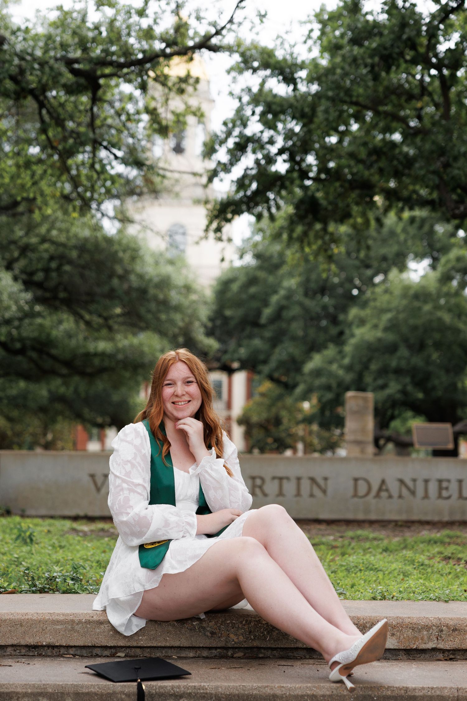 Smiling graduate in a white dress and green stole sits on stone steps near a campus sign, with a graduation cap beside her and trees behind.