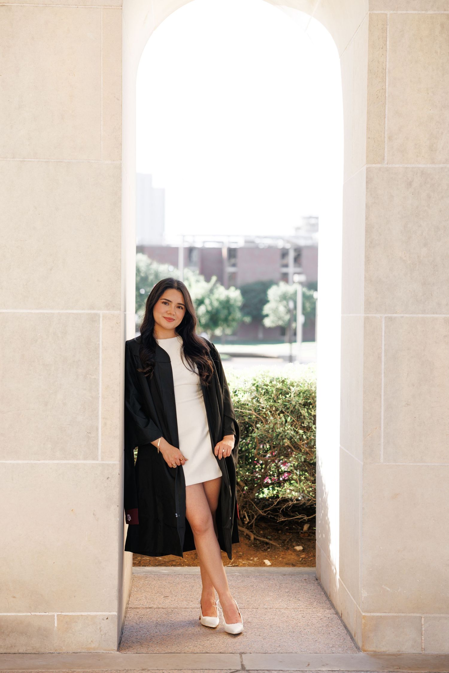 Young woman in white dress and black coat posing elegantly under a stone arch with greenery in the background.