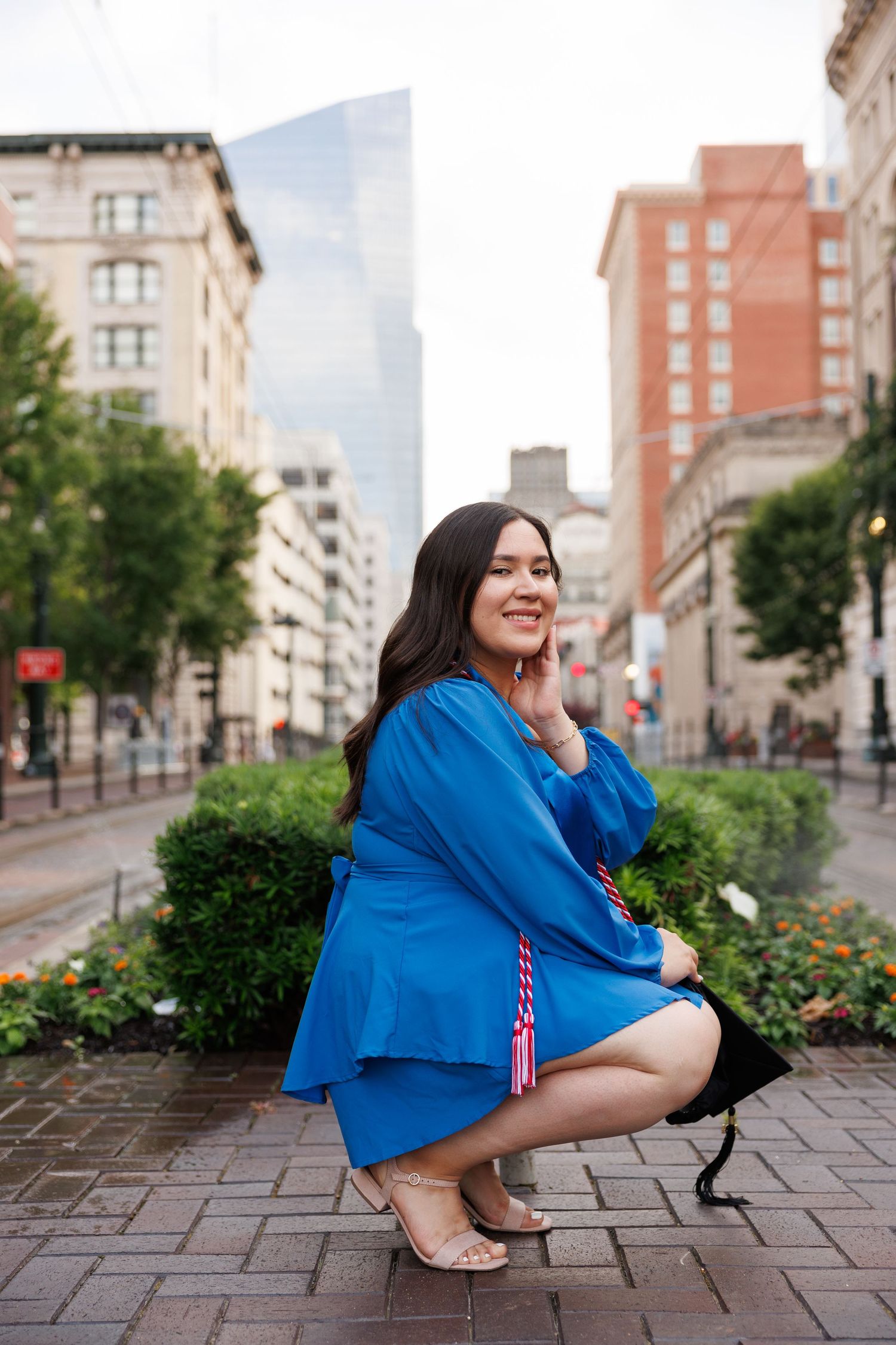 A woman in a bright blue dress crouches on a brick sidewalk in an urban setting with tall buildings in the background.
